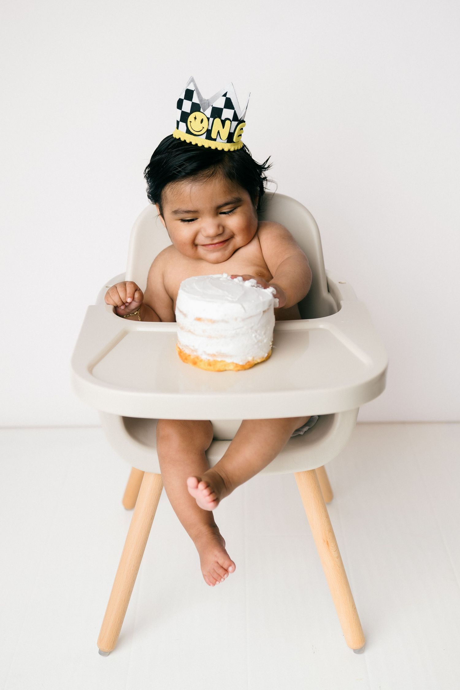 Baby wearing golden crown sits in modern white high chair with small white birthday cake on tray.