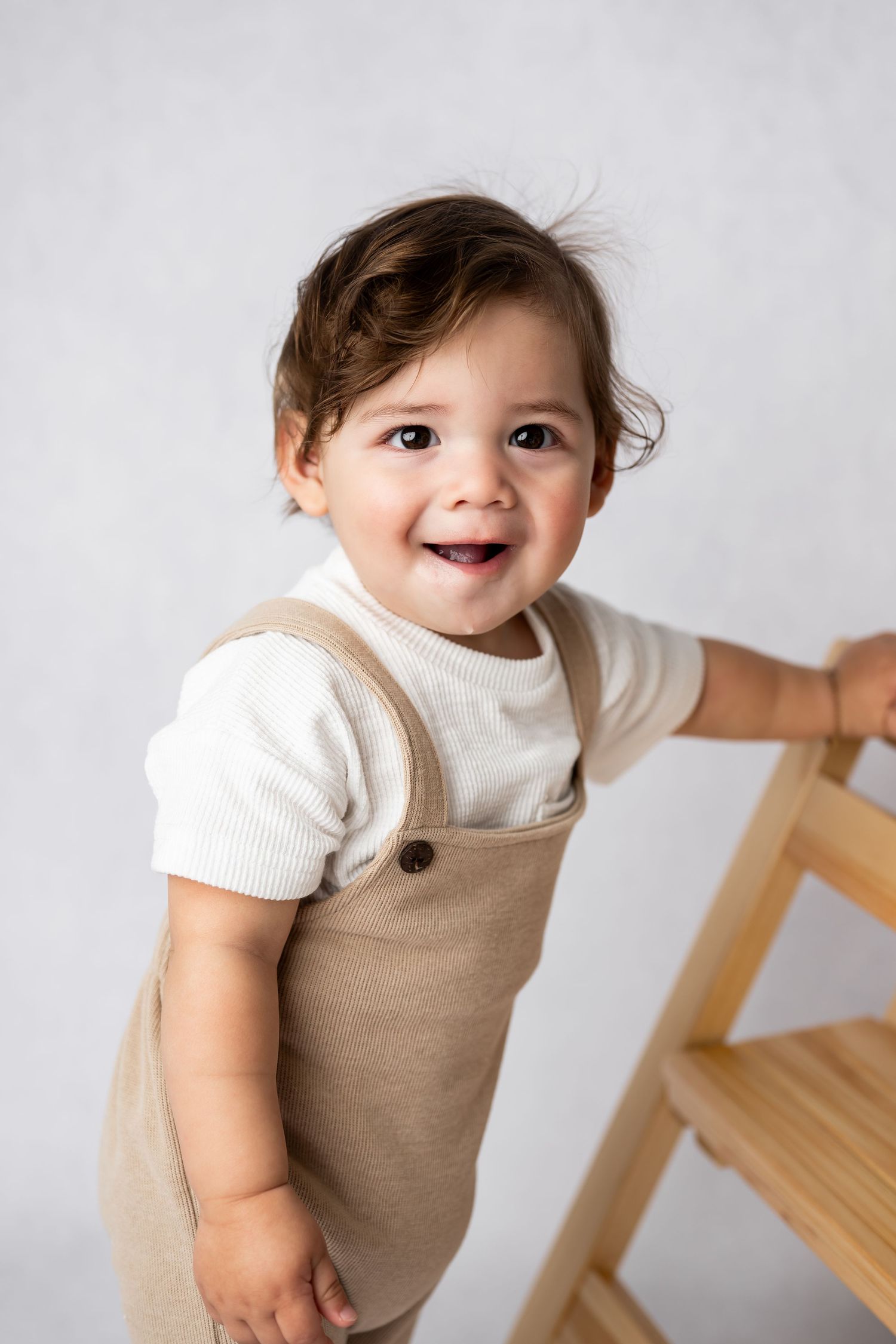 Adorable toddler in beige overalls and white shirt smiles brightly while standing near wooden furniture.