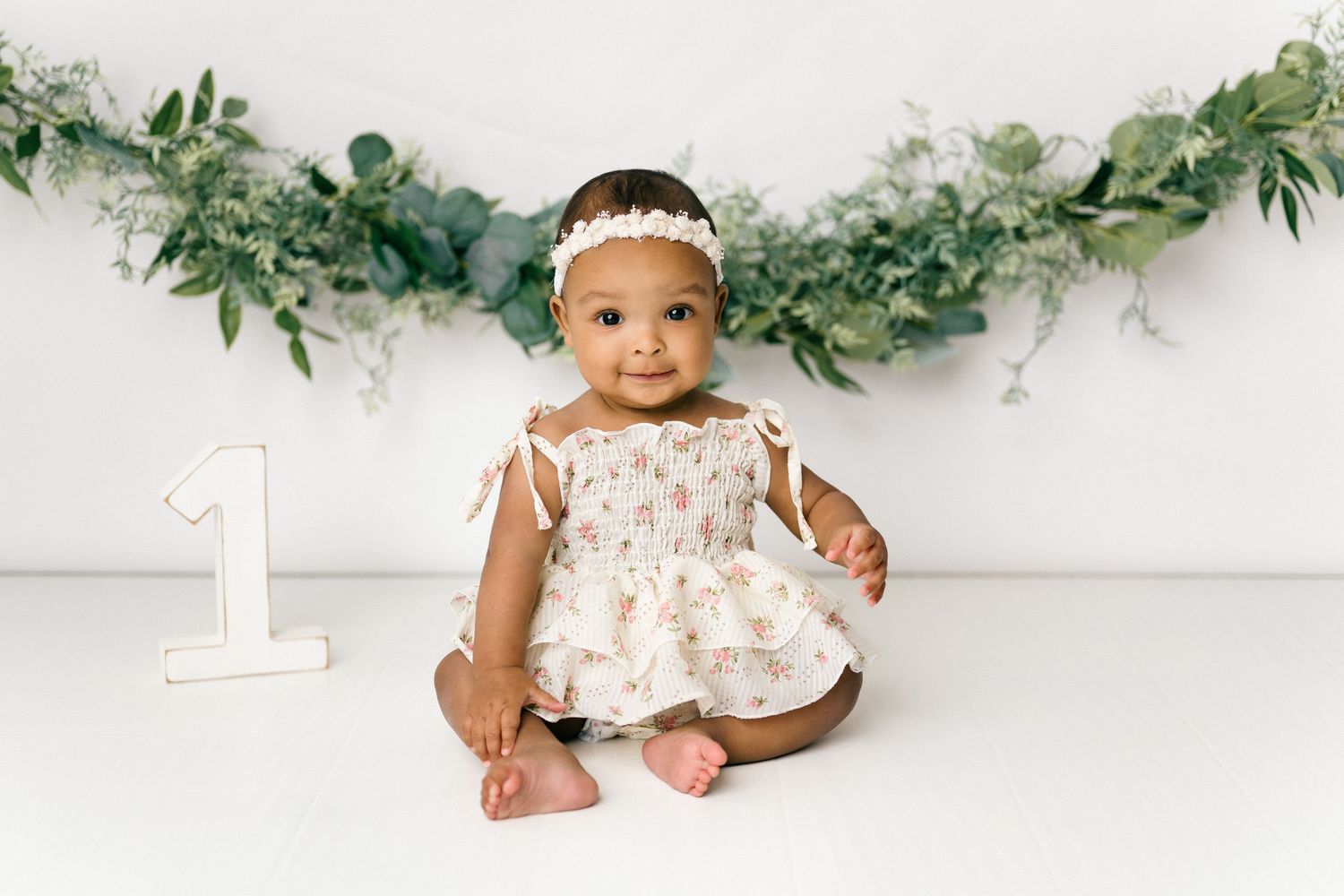 Baby in white lace dress sits on floor for first birthday photo shoot with greenery garland backdrop.