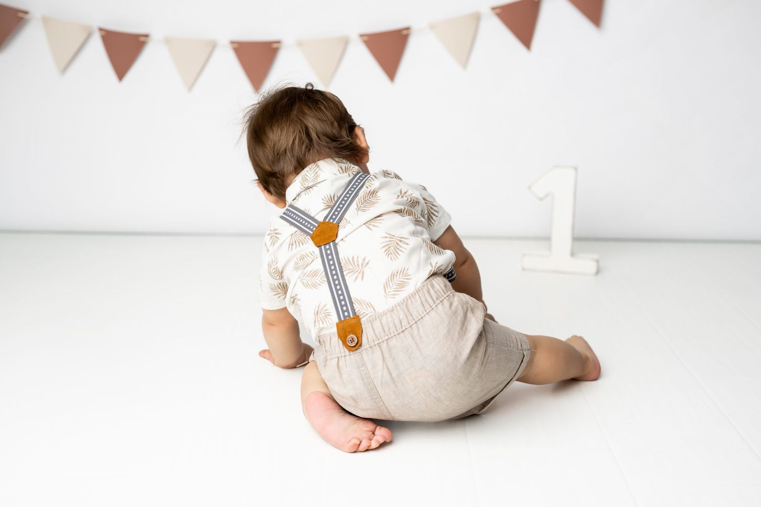 Small child sitting on floor wearing white romper with brown suspenders under festive bunting decorations.