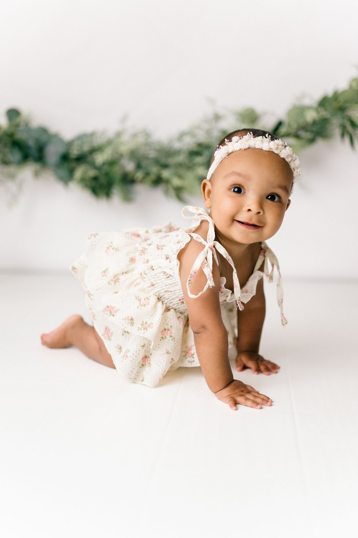 Happy baby in a white floral dress crawls on the floor with greenery garland decorations in the background.
