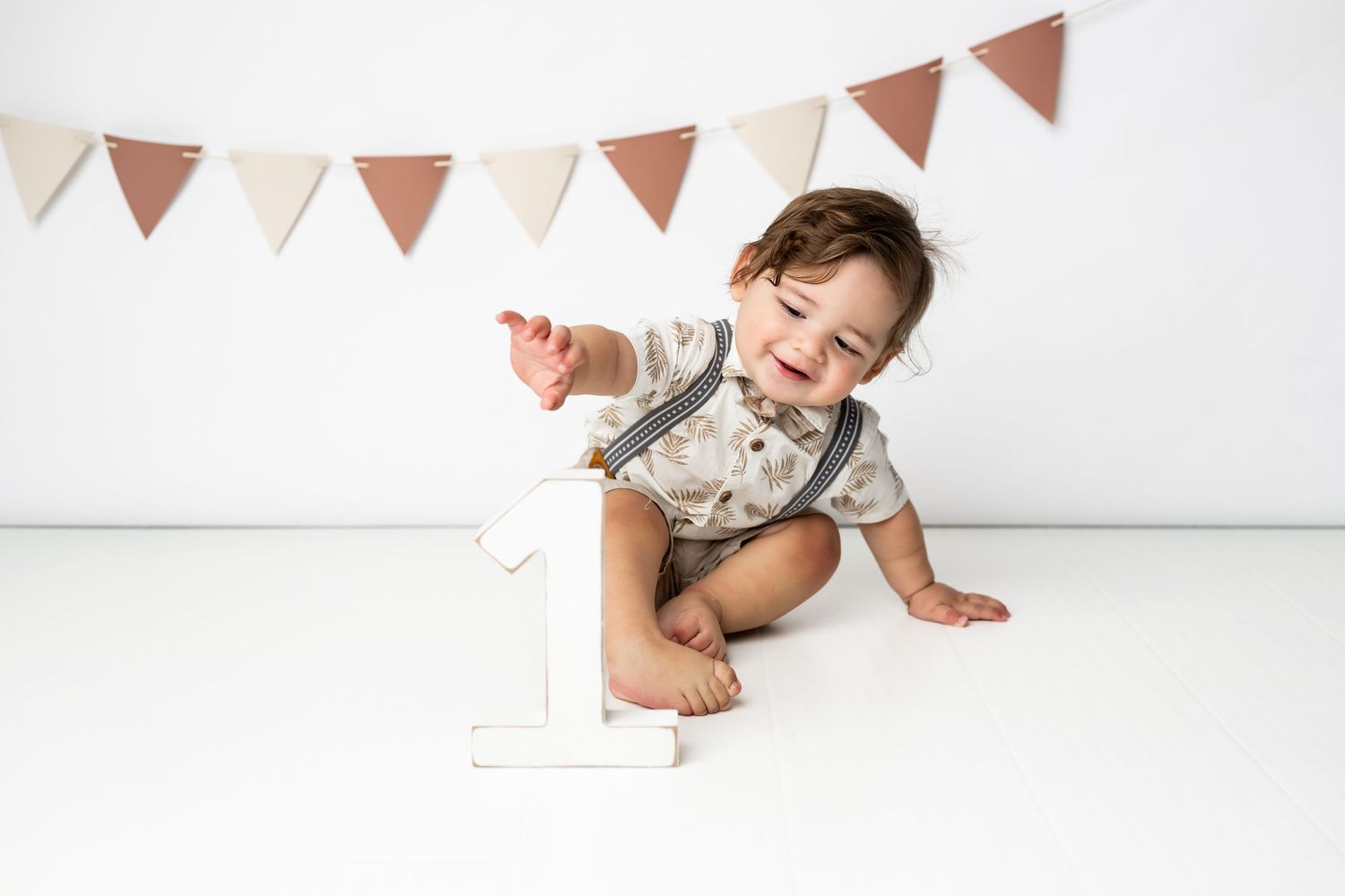 Happy baby sits next to number one decoration while celebrating first birthday under brown triangle banner garland.