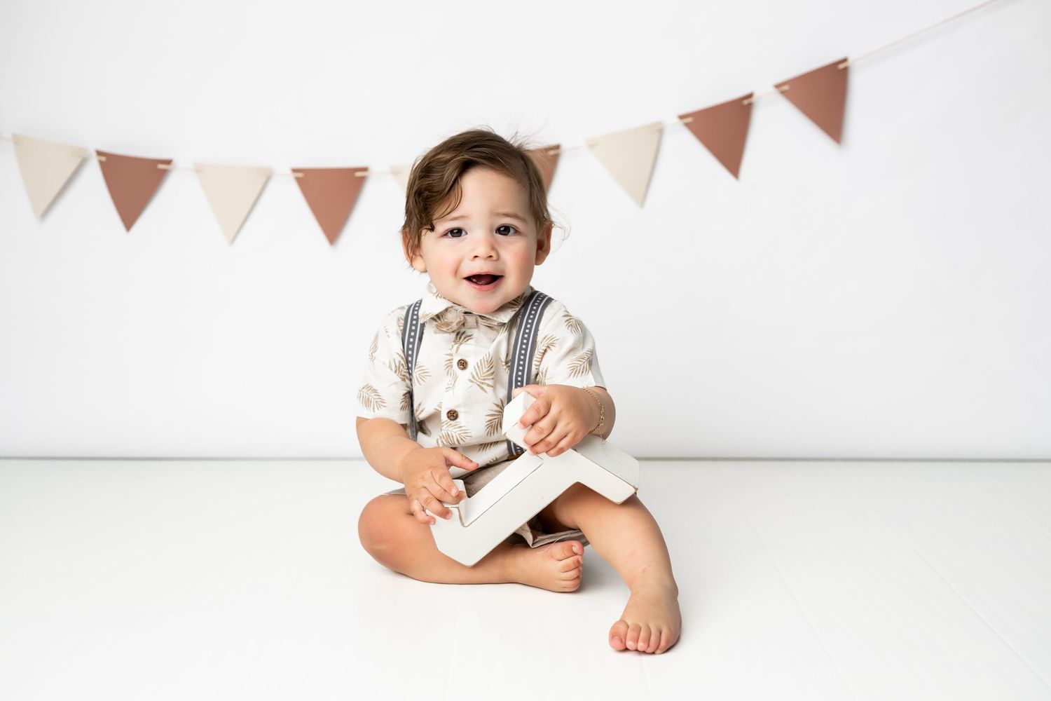 Baby in white and gray outfit sits on floor under triangle banner bunting decorations.
