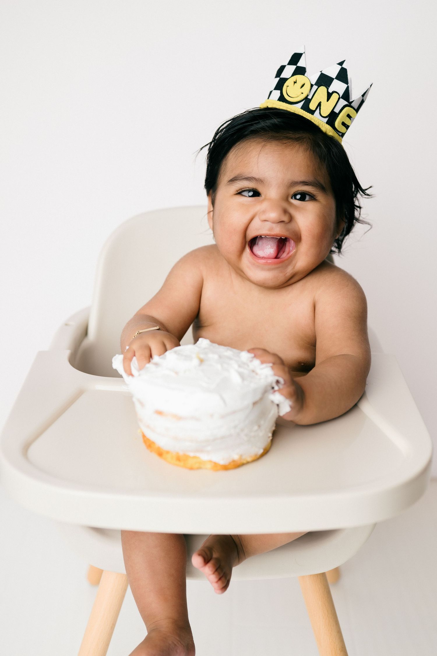 series of images showing happy baby in high chair wearing gold crown while eating cake with white frosting during first birthday.