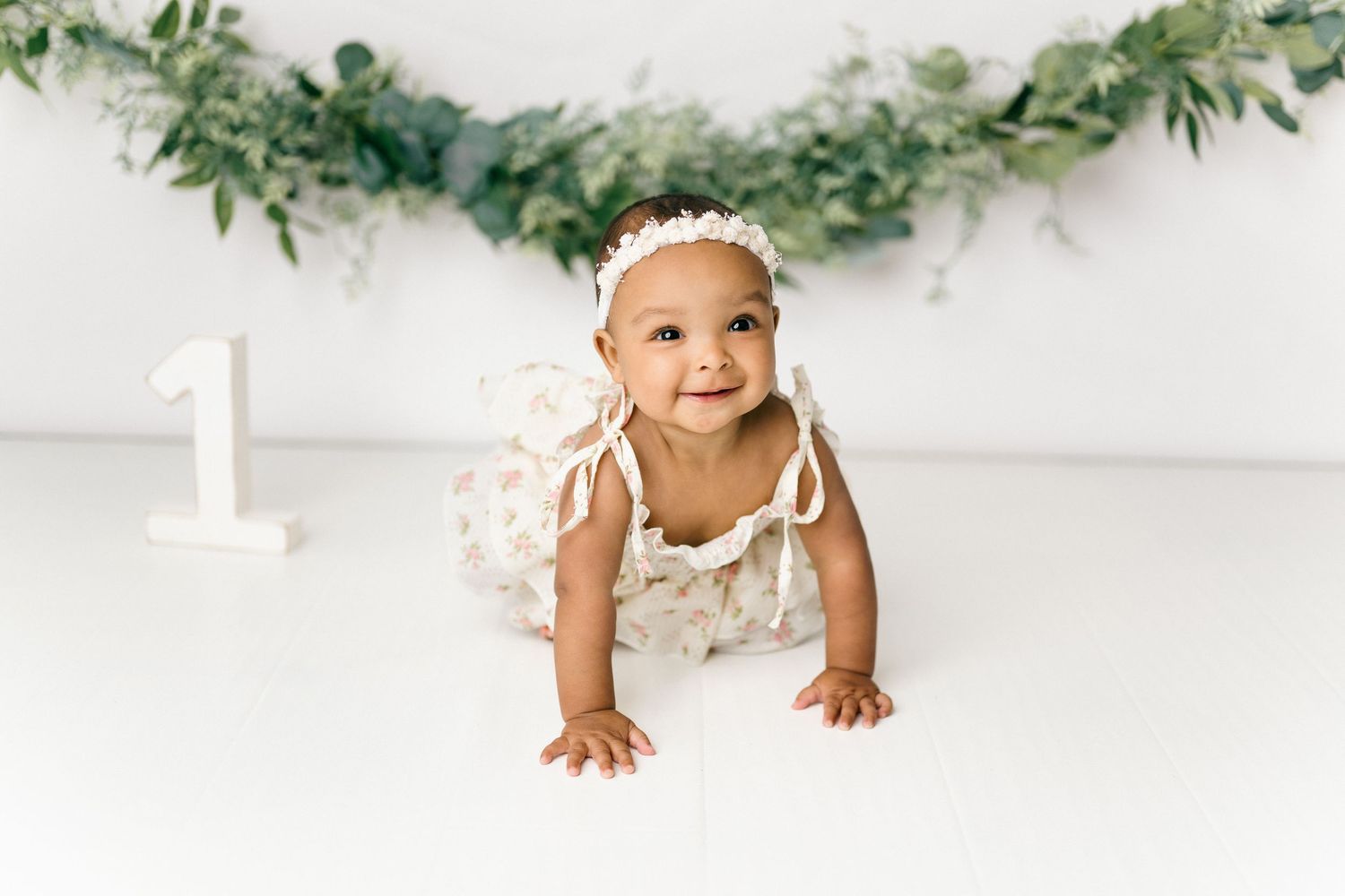 Baby in white dress crawling on floor under greenery garland during first birthday photoshoot on white backdrop.
