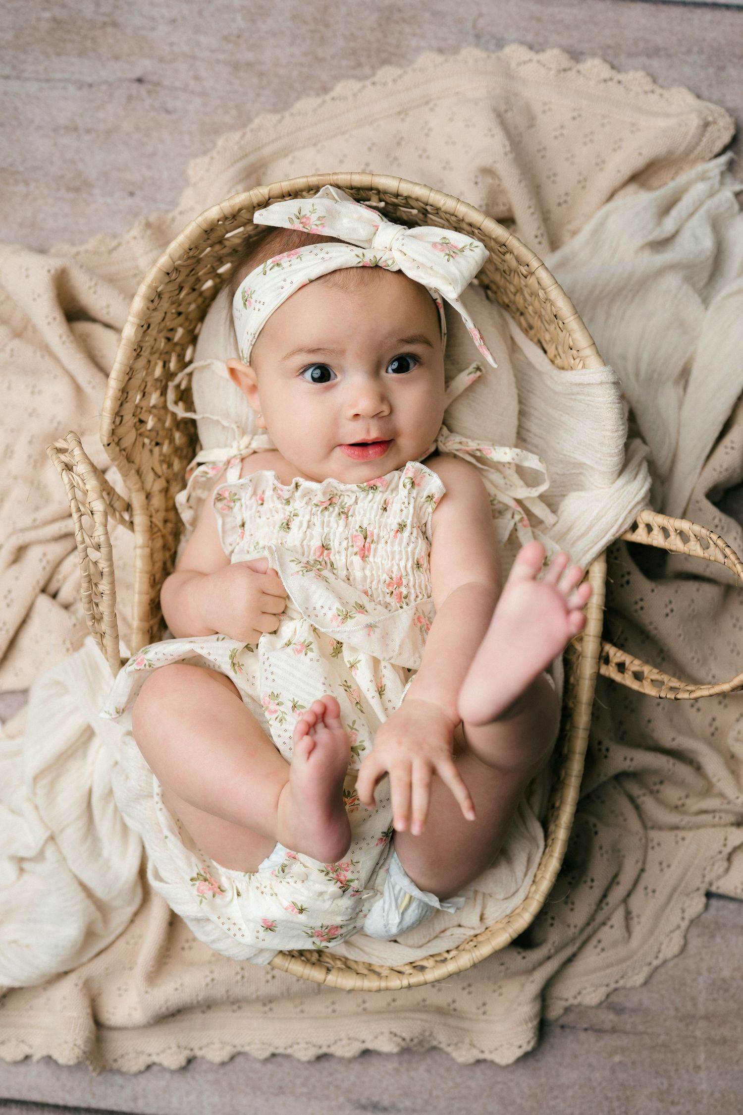 Adorable baby girl in floral romper and bow headband smiling while sitting in a wicker basket on a beige blanket.