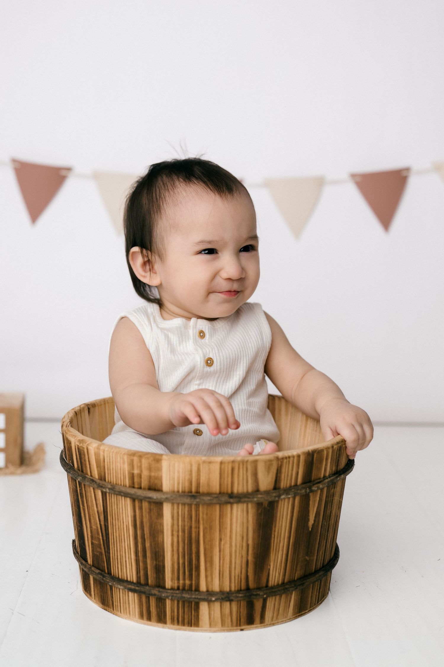 Smiling baby in white romper sitting inside a rustic wooden barrel bucket with colorful bunting flags in background.