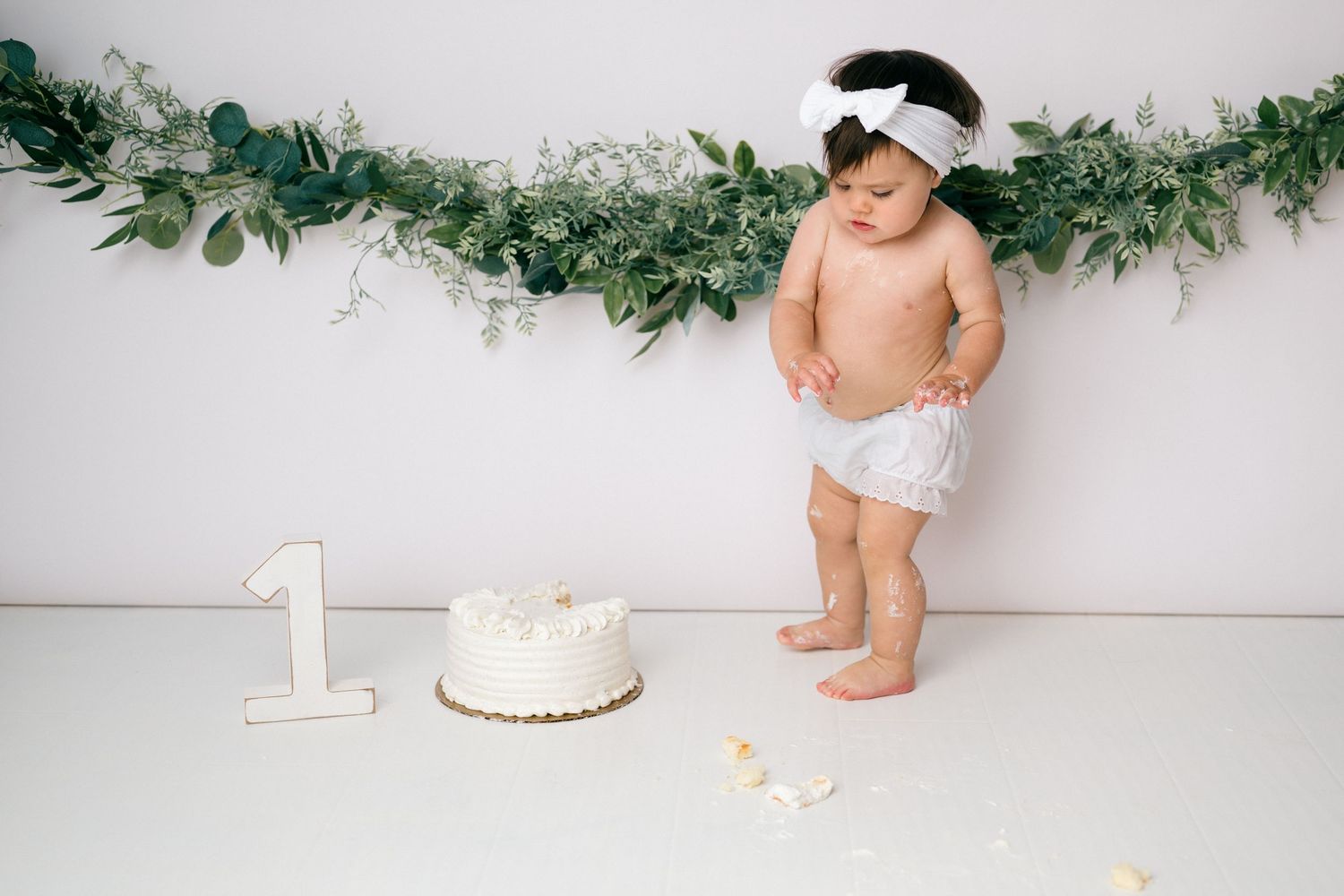 First birthday celebration setup with white cake, greenery garland and baby in diaper and bow against white backdrop.