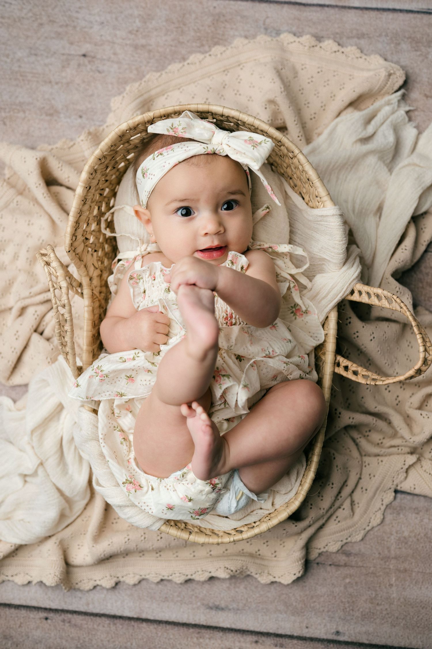 Adorable baby girl in floral romper and bow headband sitting in a wicker basket on a beige knit blanket.
