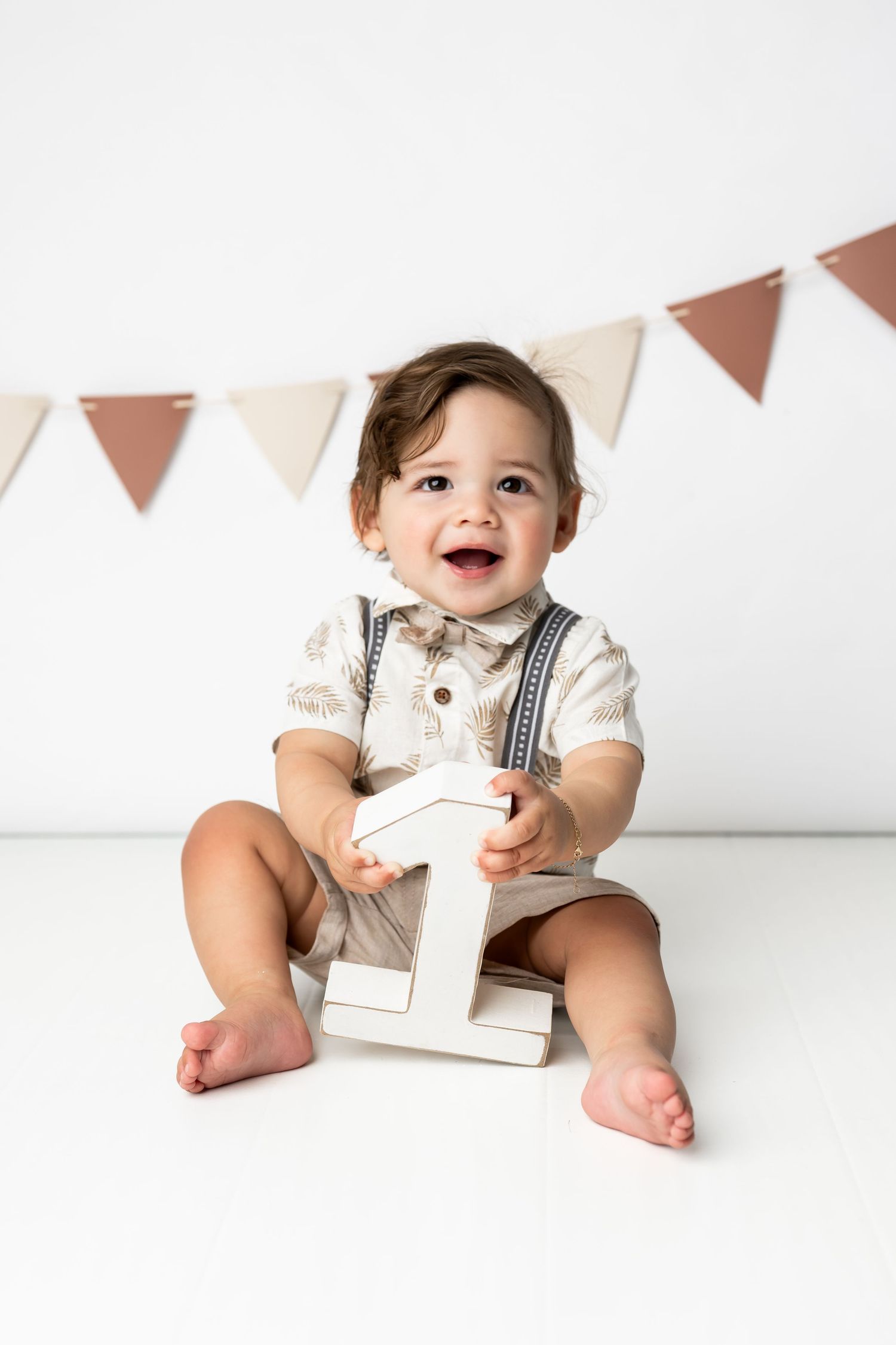 Happy baby in white shirt and overalls sits on white floor holding number one decoration under triangle party banner.