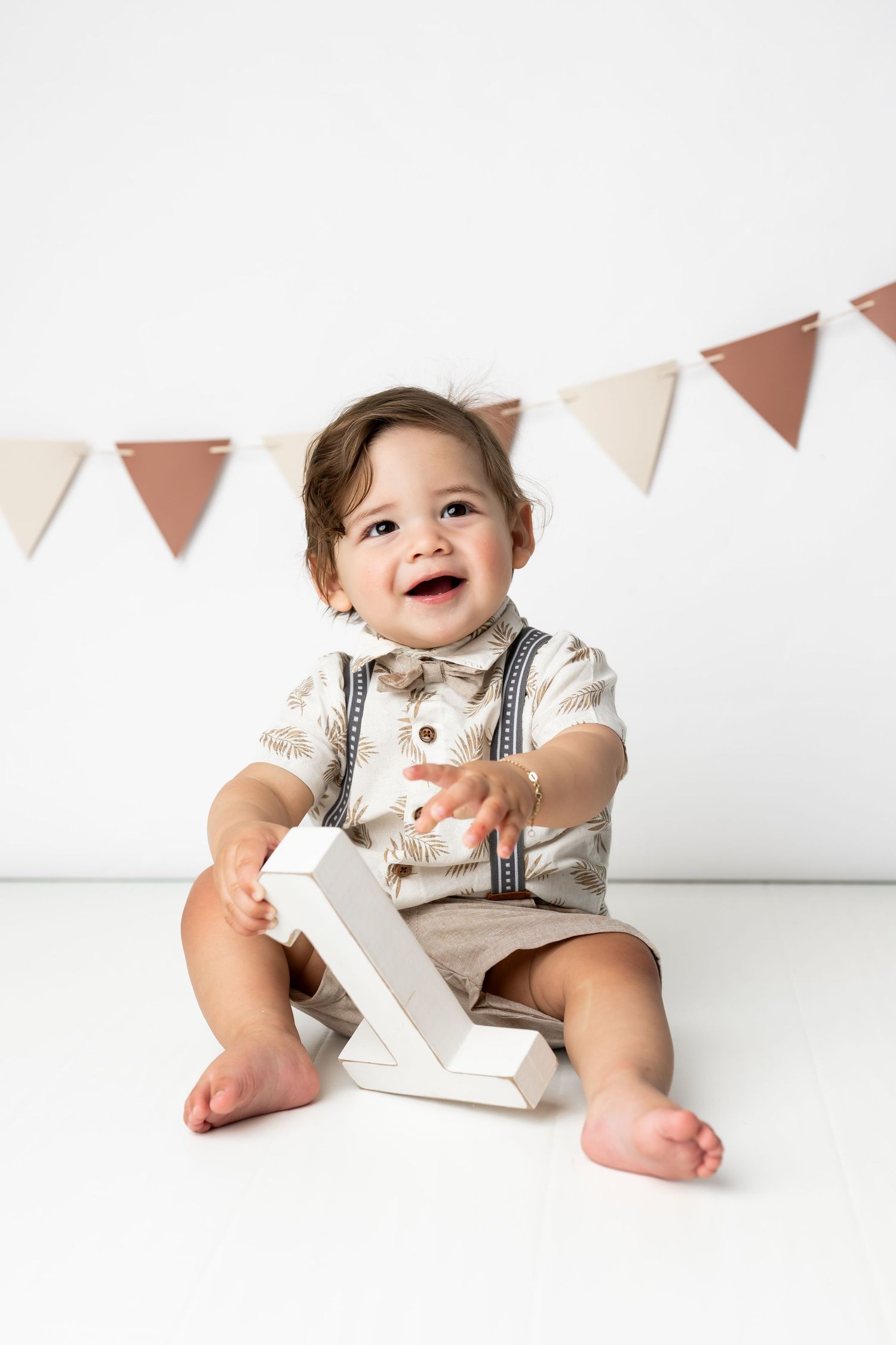 A smiling baby wearing suspenders and formal attire sits on a white background with festive pennant decorations.