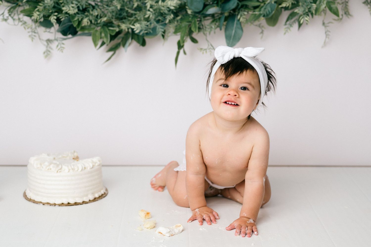 A collage of 6 images showing a baby's first birthday cake smash photo session with white backdrop and greenery decorations.