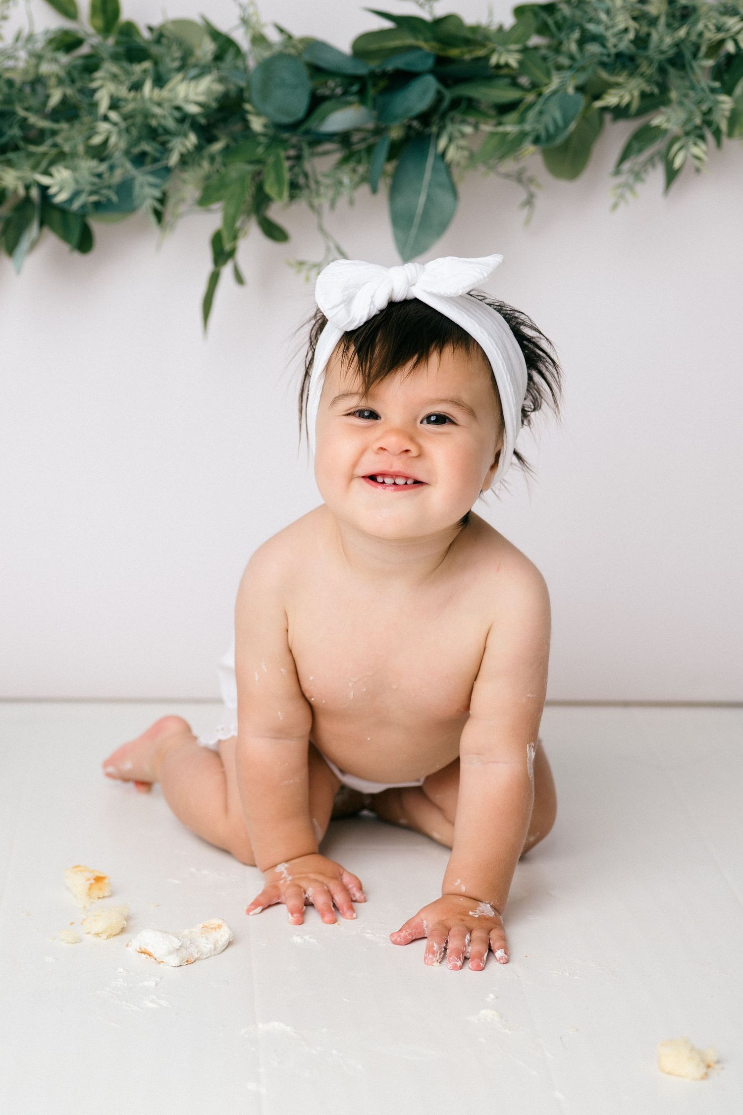 A baby wearing leaves and greenery while crawling on white floor with scattered orange flower petals.