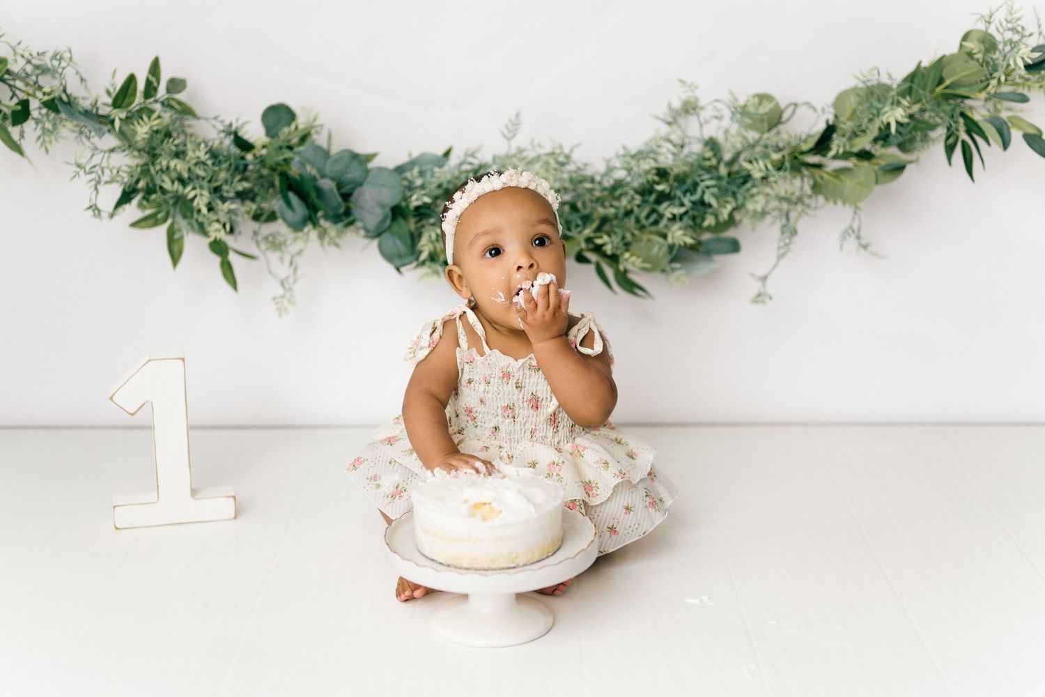 Baby in floral dress sits near white number one decoration and eucalyptus garland for first birthday cake smash photo.