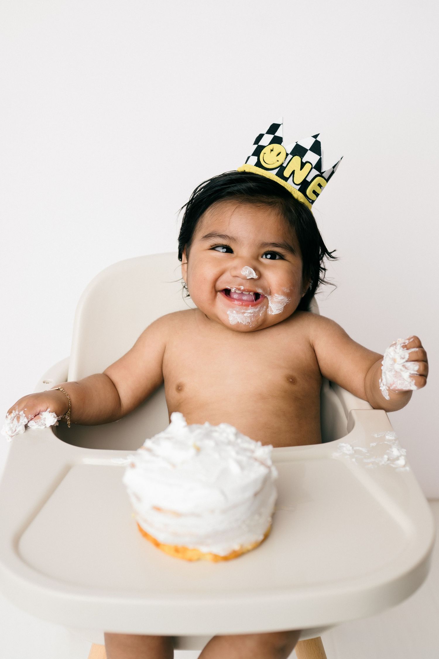 A baby with a birthday crown plays with a white frosted cake in a high chair, getting messy with the dessert.