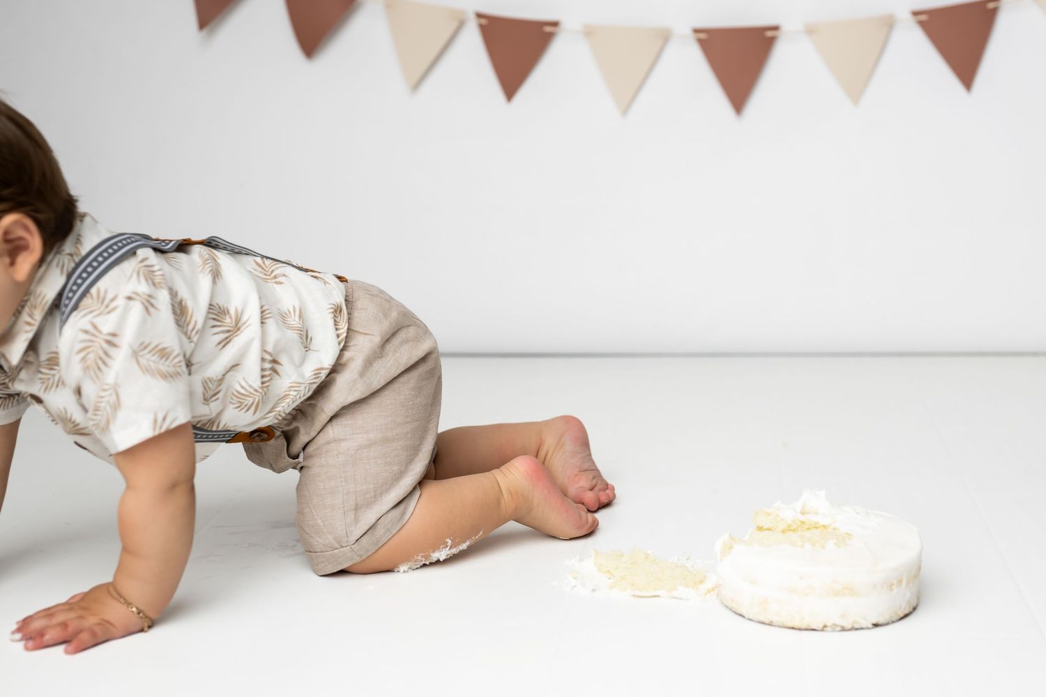 Small child in light romper crawling towards white birthday cake with bunting banner hanging on wall above.