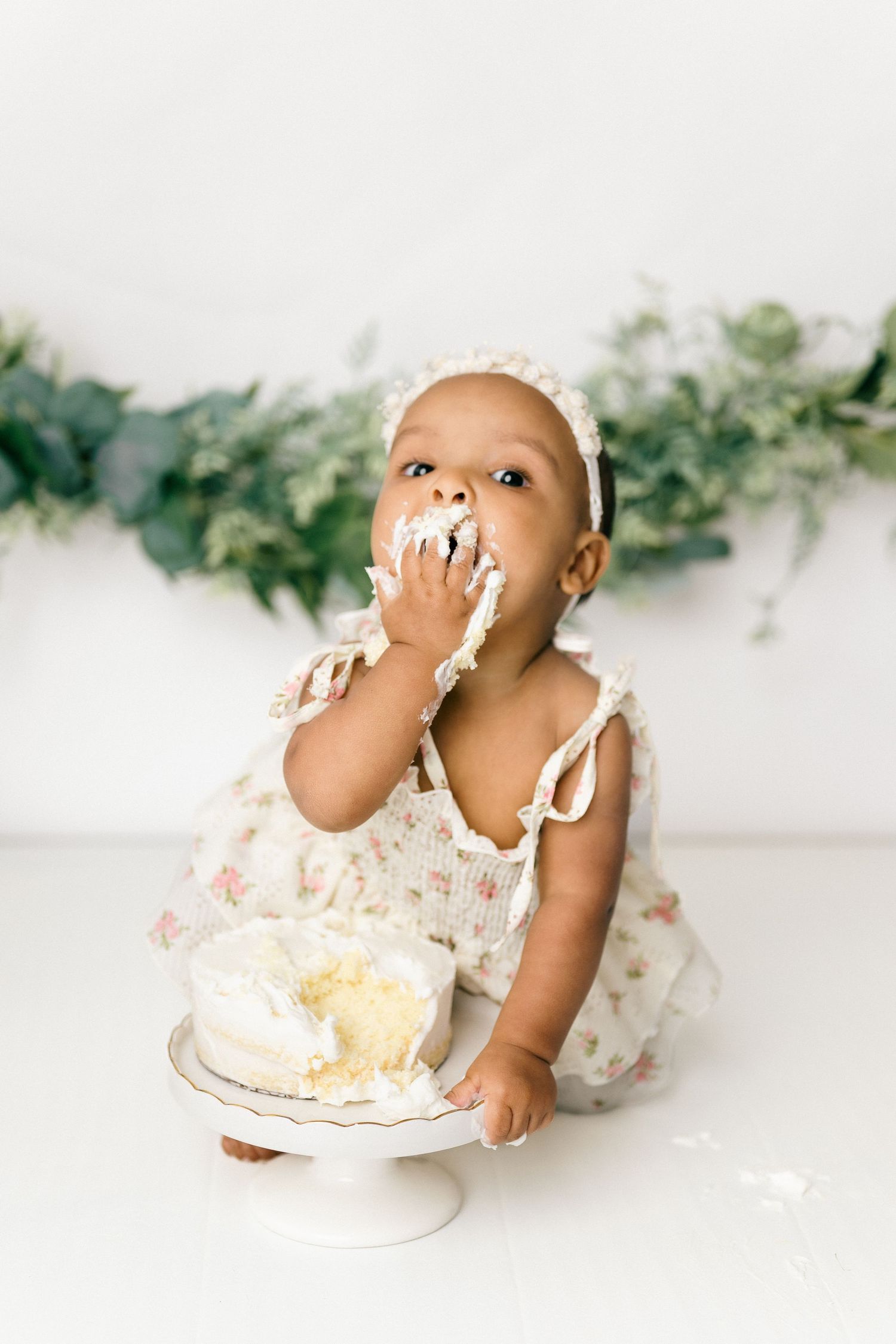 Little one sitting with a smashed cake wearing a floral dress during a cake smash photo session.