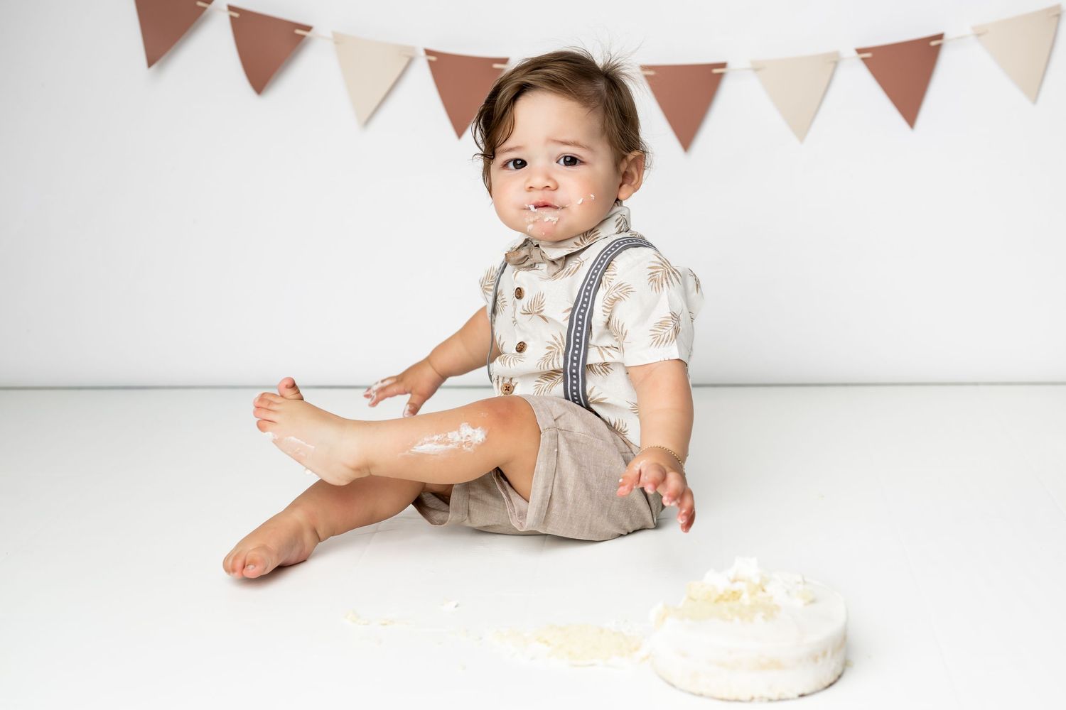 A toddler in vintage-style outfit with suspenders sits barefoot on white floor near decorative bunting and cake.