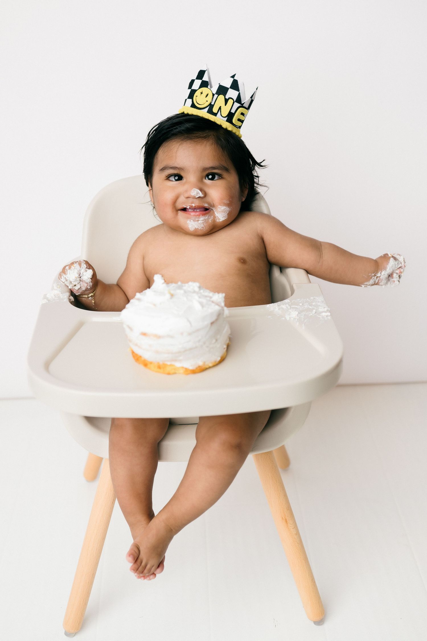 Happy baby wearing a crown sits in white high chair with smash cake during first birthday photo session.