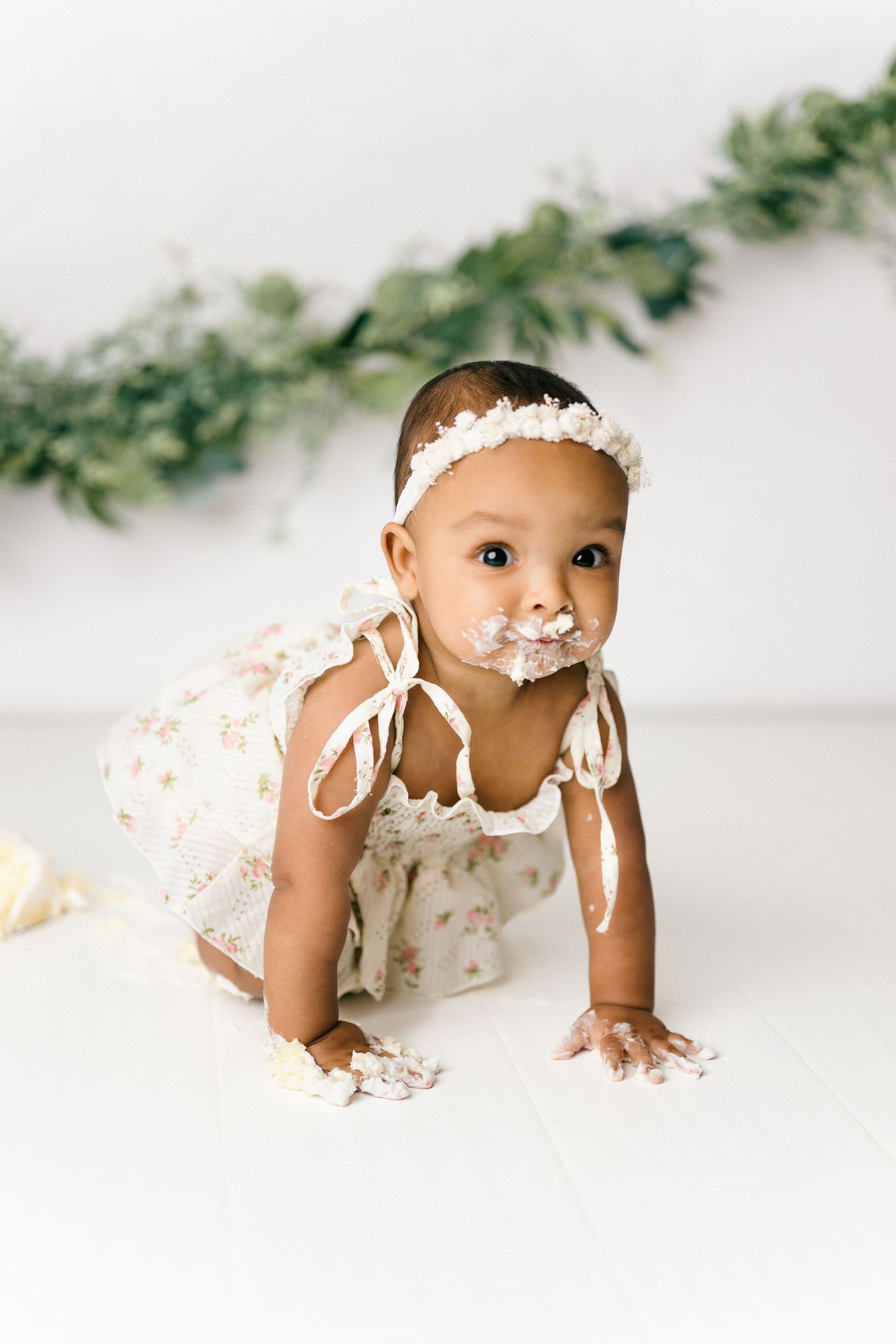 Baby in white lace dress with white headband crawls on floor while covered in cake frosting during smash cake photoshoot.