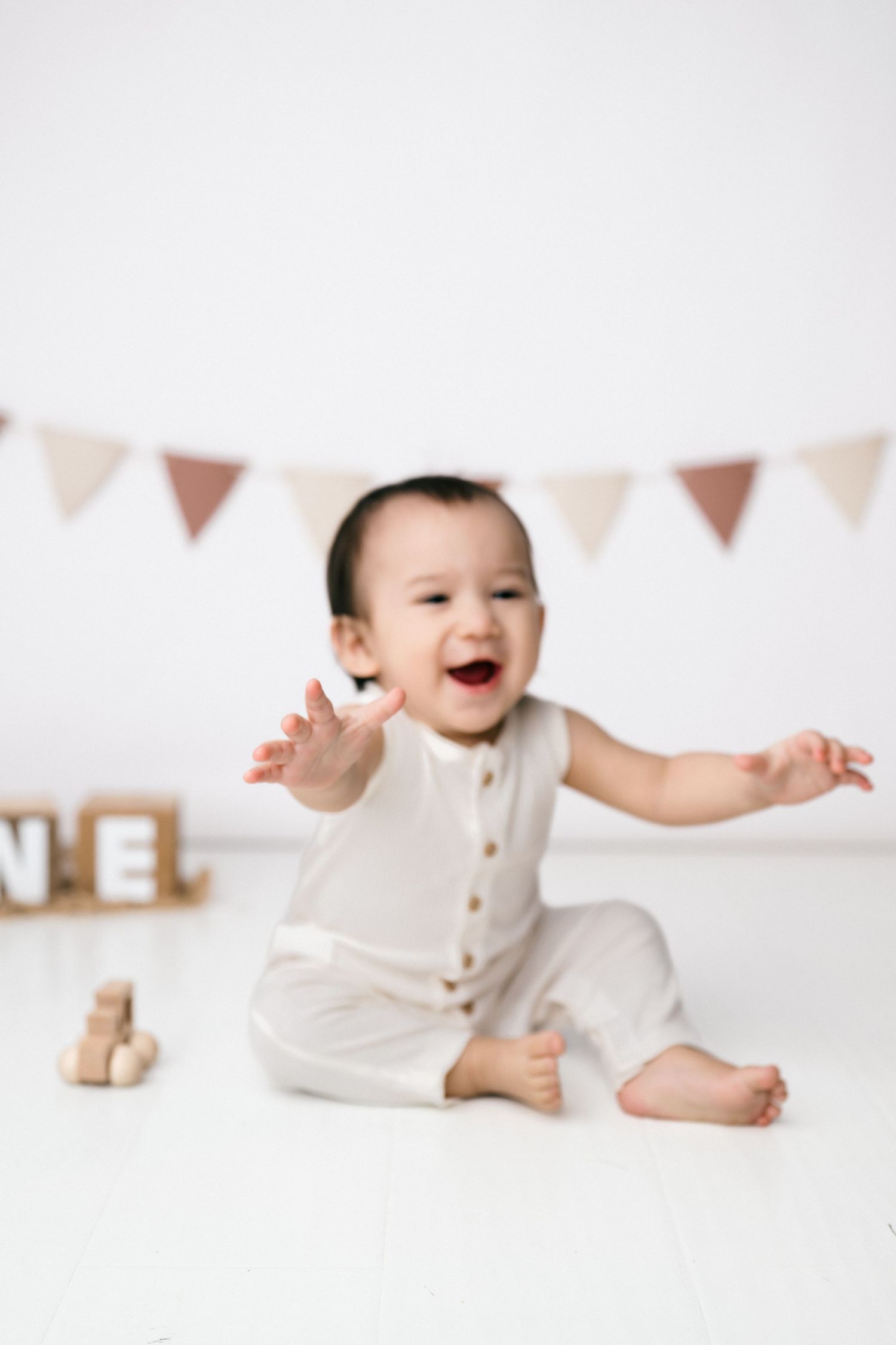 A joyful baby in a white romper sits on a white floor, arms outstretched, laughing, with bunting and wooden toys behind.