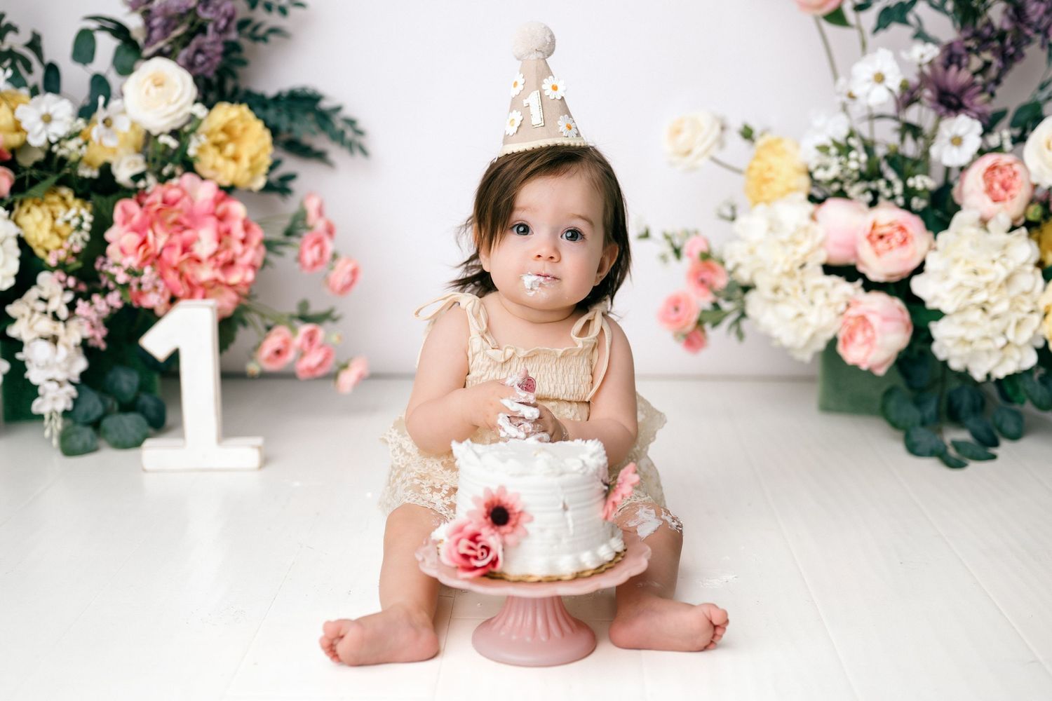 Baby girl in gold romper and crown smashes first birthday cake surrounded by pink and white flowers.