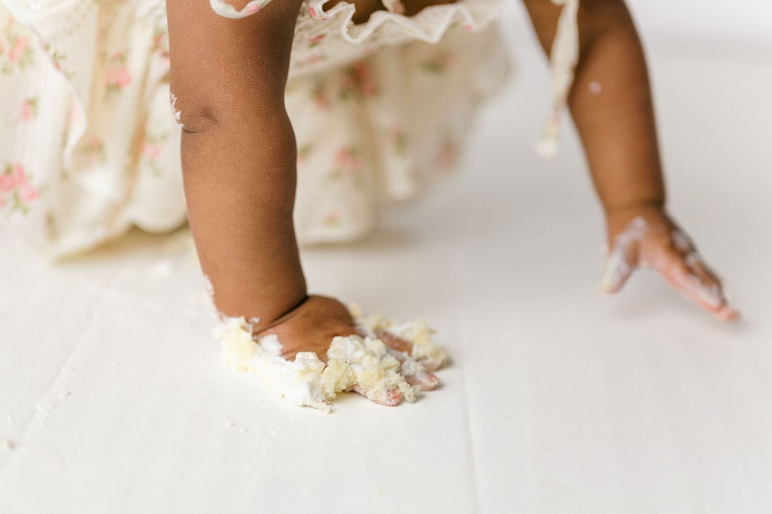 Small hands reach down to smash white frosting and cake on a white surface in a cake smash photo session.