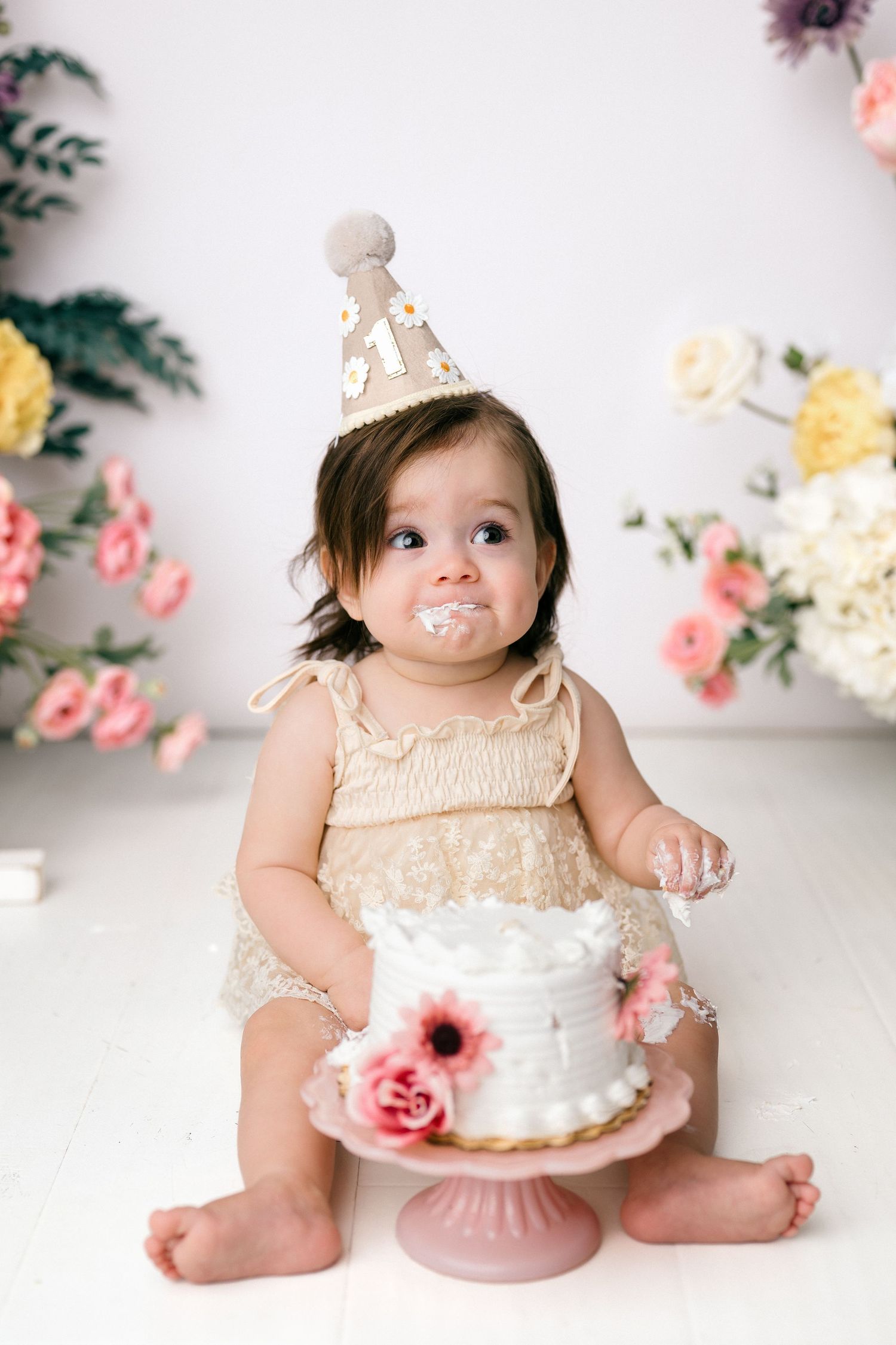 Baby girl in lace romper and birthday hat smashes white floral cake on pink stand during first birthday photo session.
