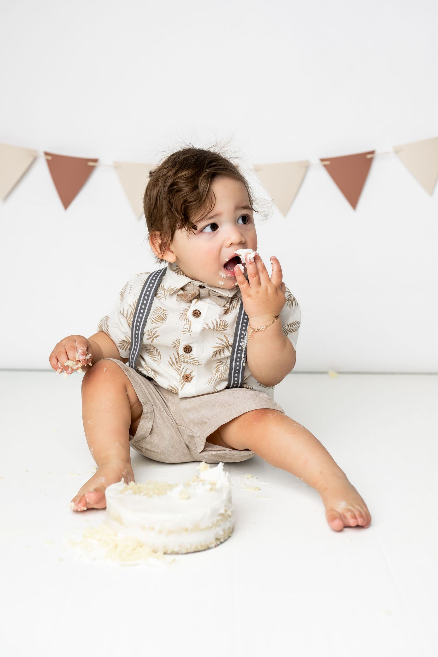 Baby wearing suspenders and white shirt sits enjoying vanilla cake at first birthday celebration with bunting banner.
