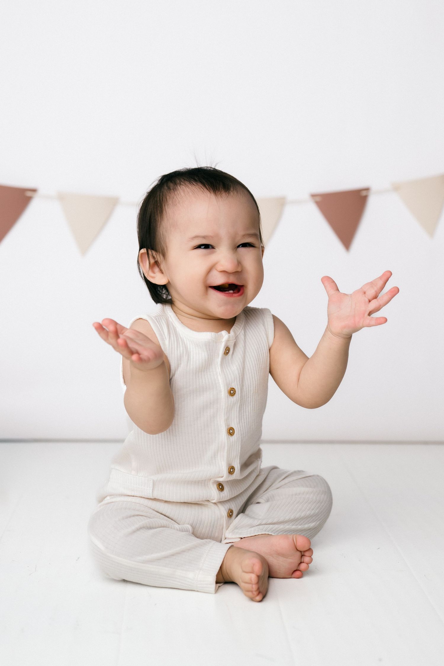 A joyful baby in a cream romper sits cross-legged, clapping and laughing, with festive bunting flags in the background.