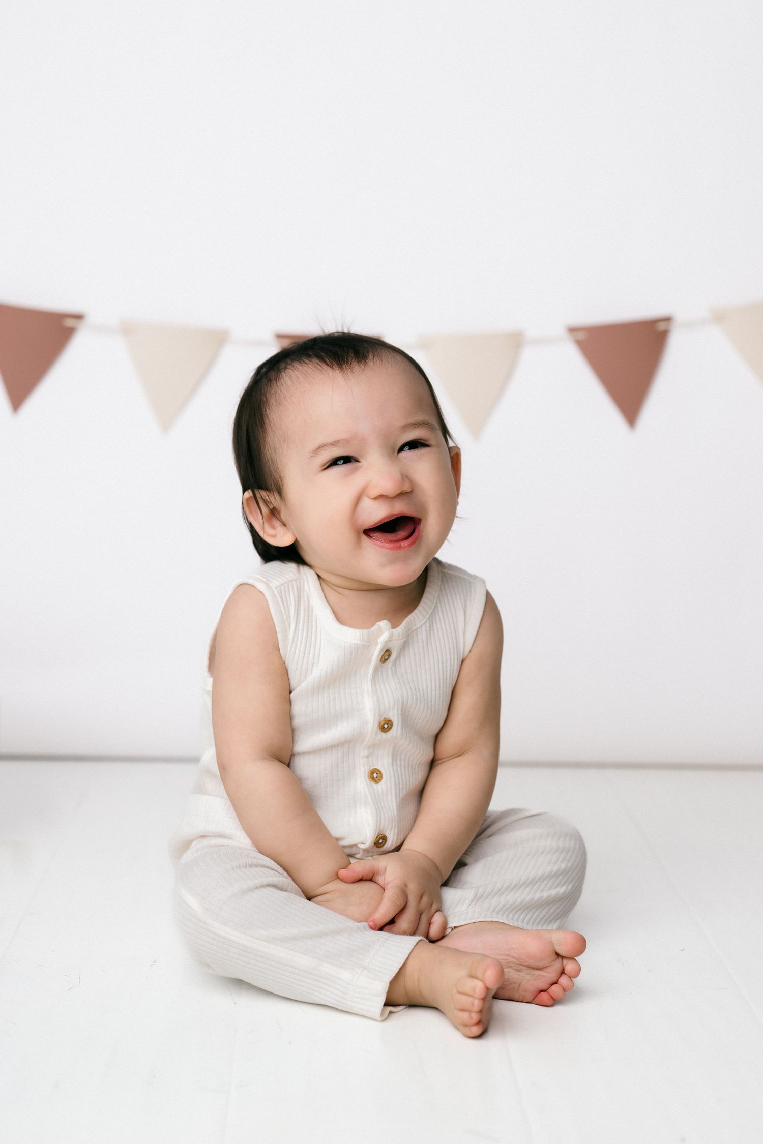 A happy baby in a cream outfit sits cross-legged on a white floor, laughing joyfully with bunting flags in the background.