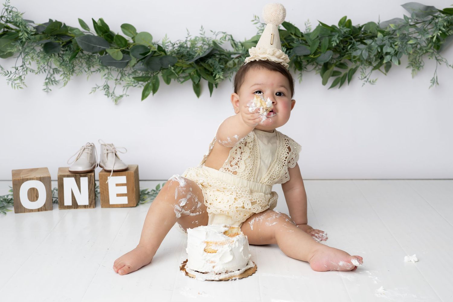 Baby wearing white outfit sits on floor next to smashed birthday cake during first birthday cake smash photo session.