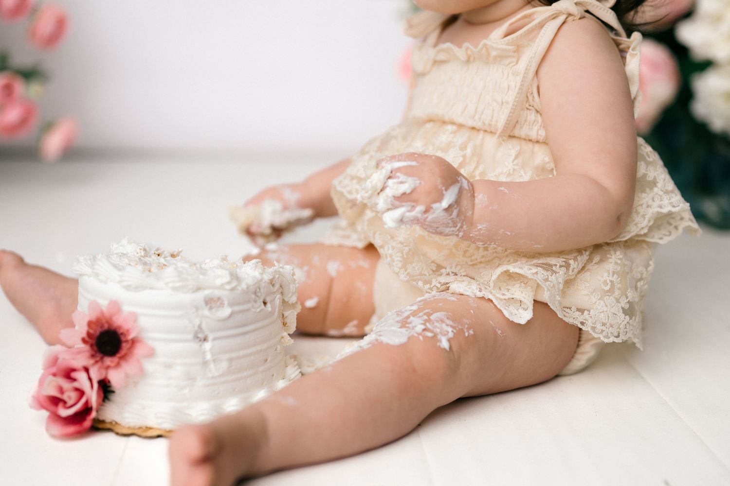 Baby girl in lace romper smashing white cake during first birthday cake smash photo session.