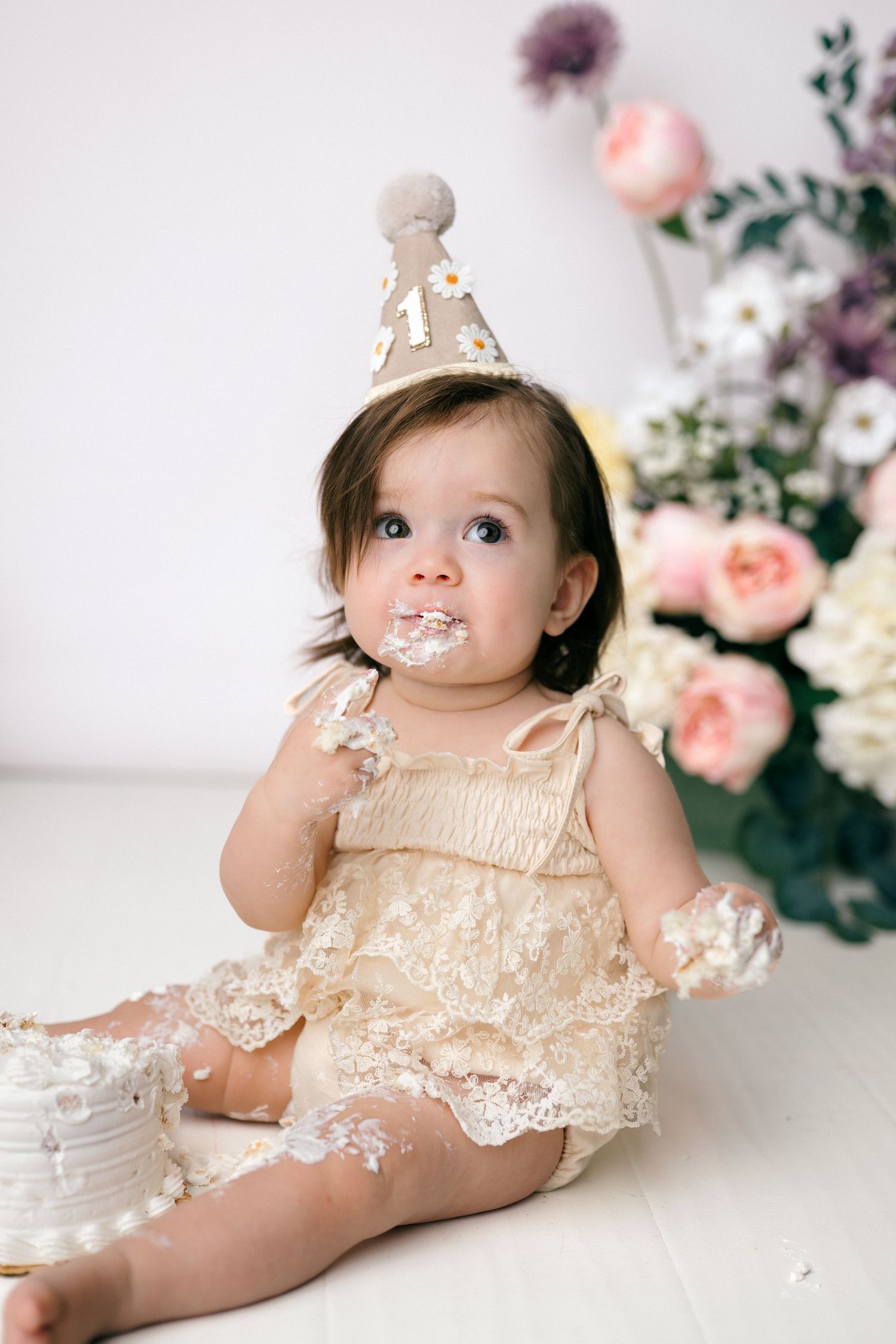 Baby girl in lace romper and birthday hat smashes first cake, sitting on white floor with flowers.