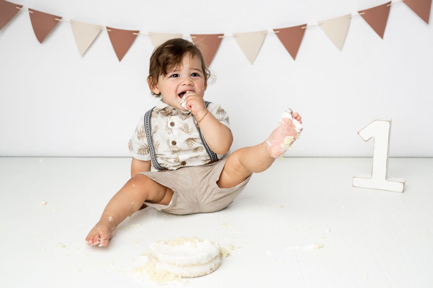 Baby boy in suspenders and bow tie sits with birthday cake and banner during first birthday photoshoot celebration.