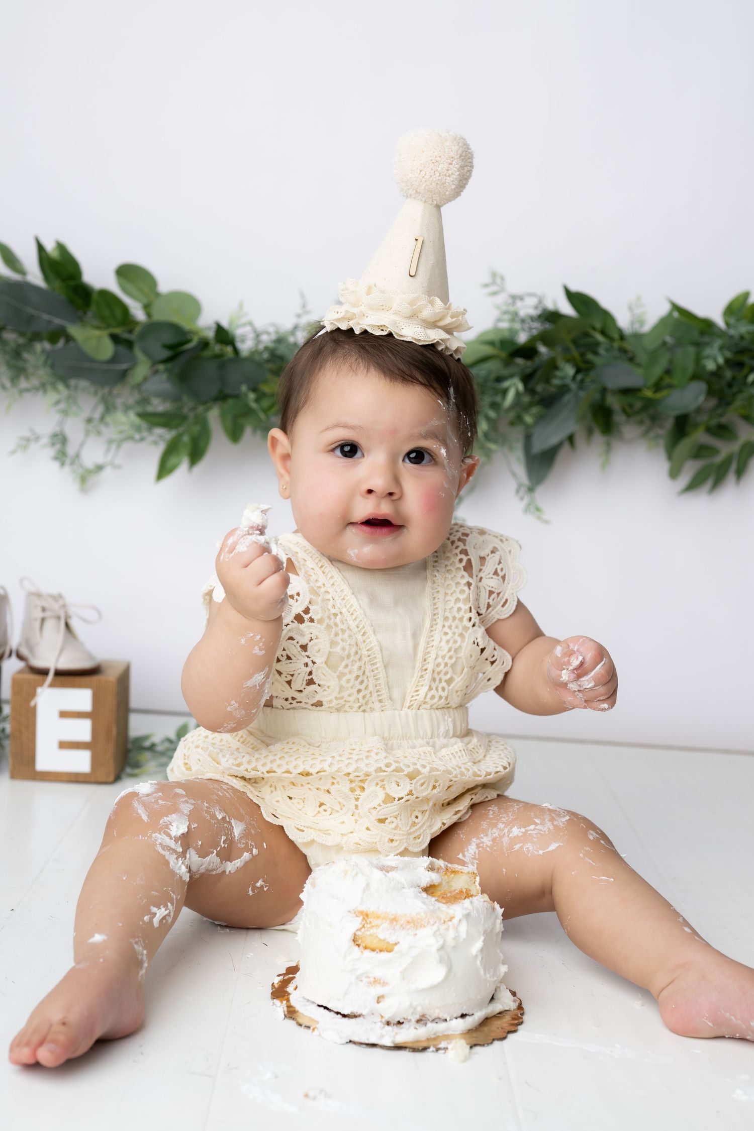 Baby in cream lace dress enjoys first birthday cake smash photo session with white cake and greenery backdrop.