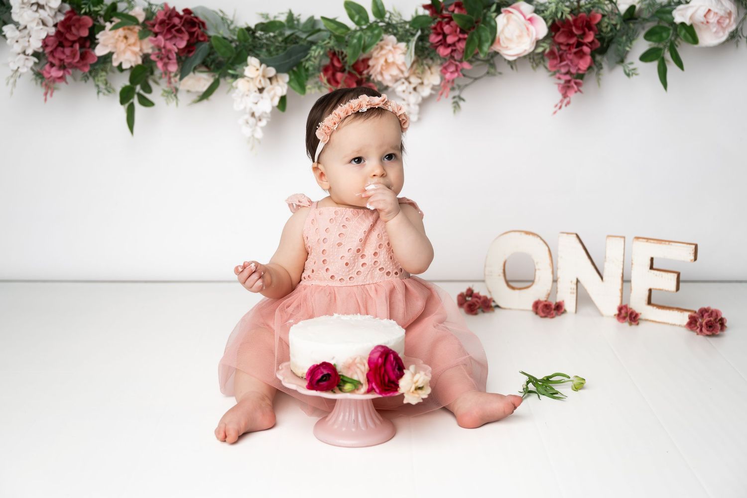 First birthday celebration setup with pink dress, cake smash, floral garland, and wooden 'ONE' letters against white backdrop.