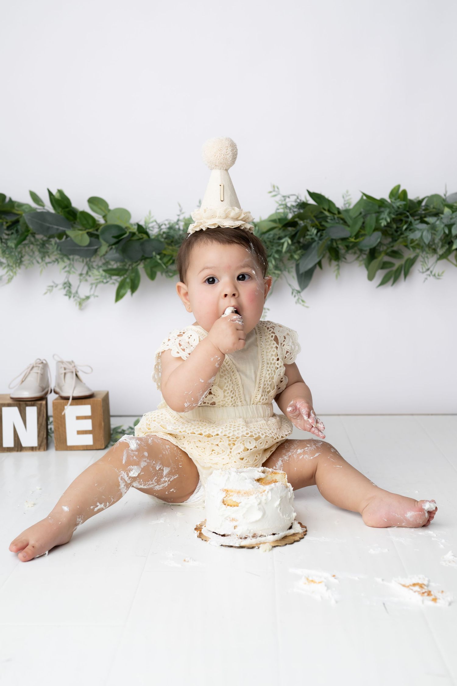 Baby in cream lace dress enjoying a cake smash photo session with greenery garland backdrop.