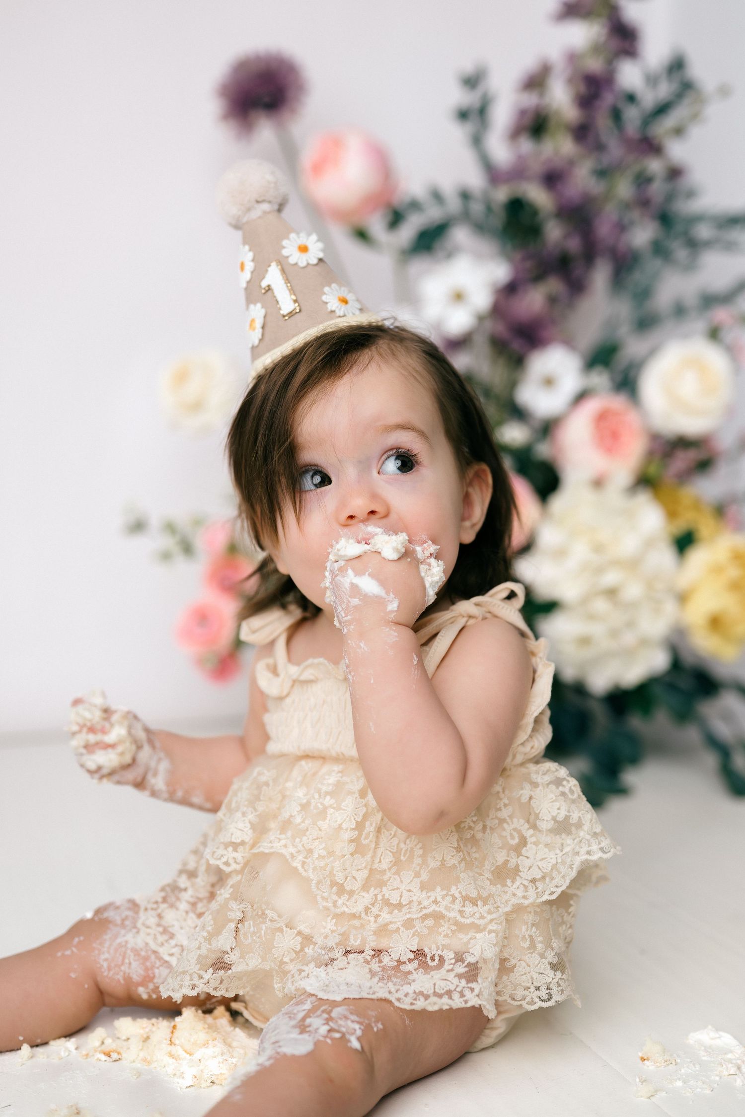 Adorable baby girl in lace romper eats birthday cake with frosting on her face during cake smash photo session.