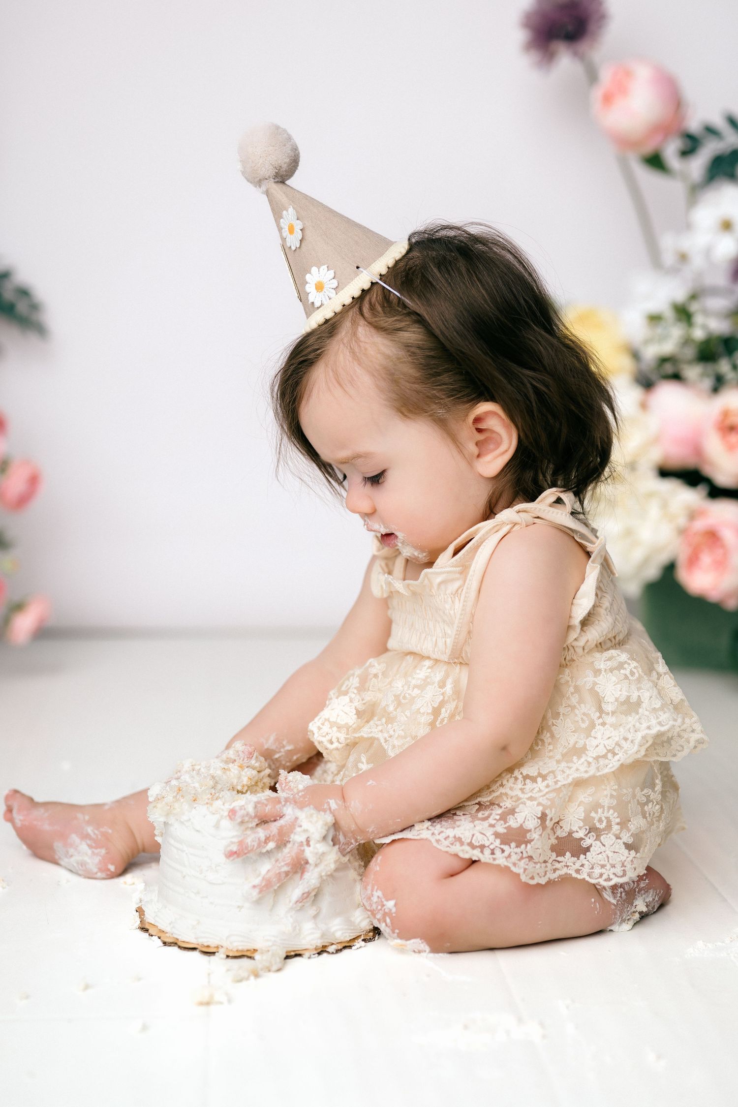 Baby girl in lace dress and birthday hat smashing her first birthday cake on white background.