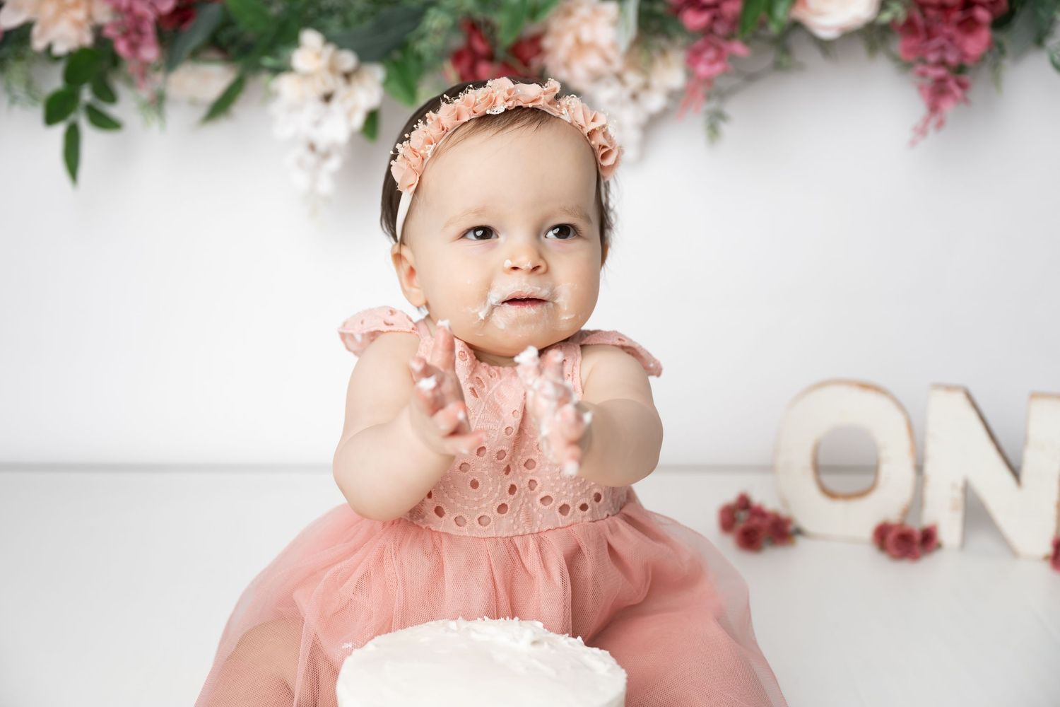 Decorative pink floral backdrop with white cake and 'ONE' text for milestone celebration photography session.