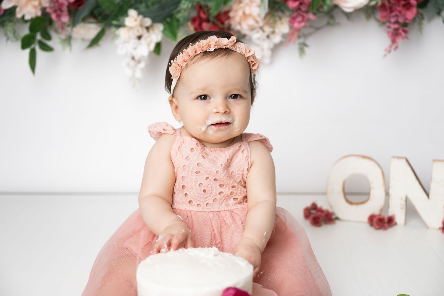 A small figure in a pink dress and floral headband sits with a white cake against a floral backdrop with decorative letters.