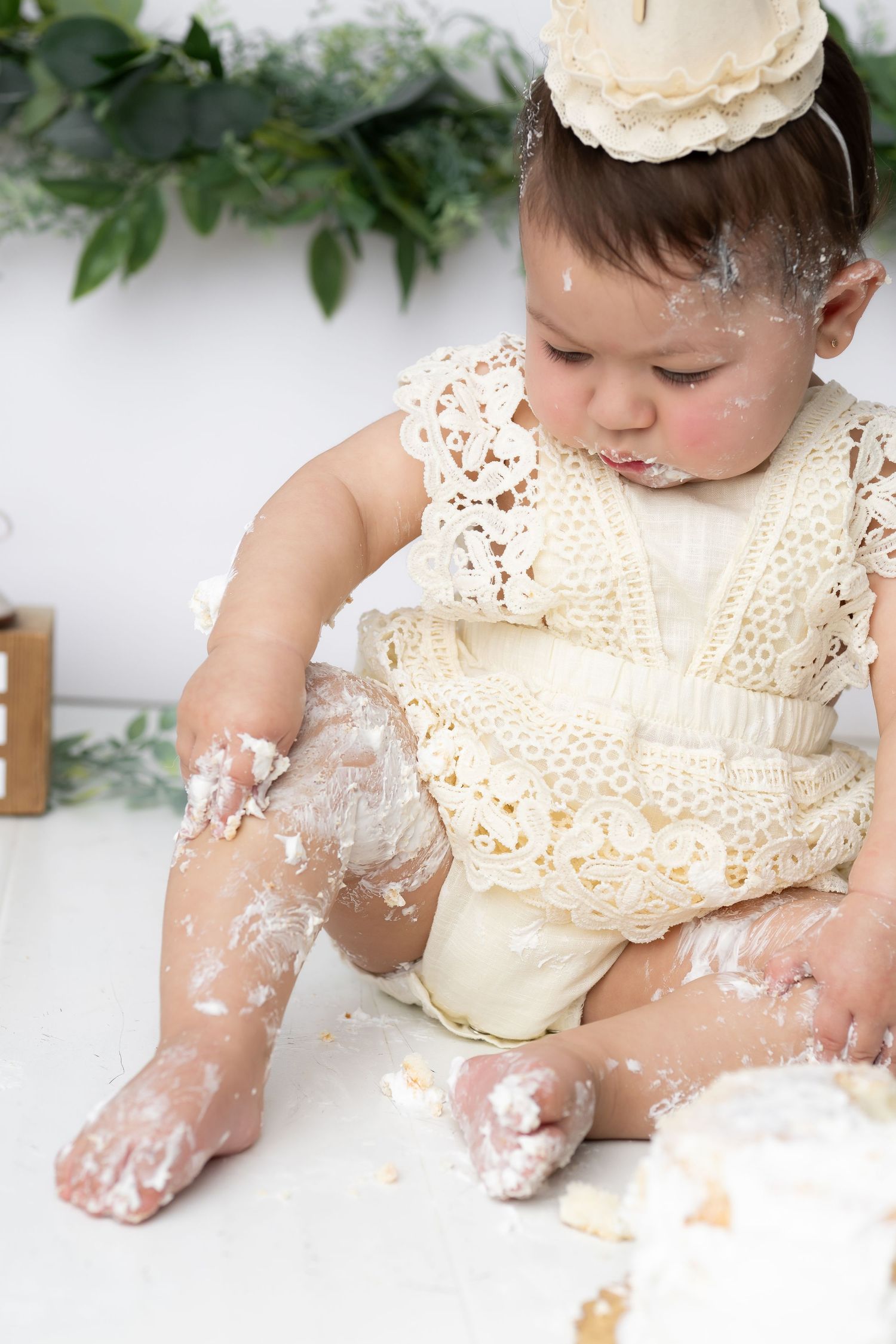 Baby in vintage-style dress getting messy with cake during milestone photo session against white backdrop.