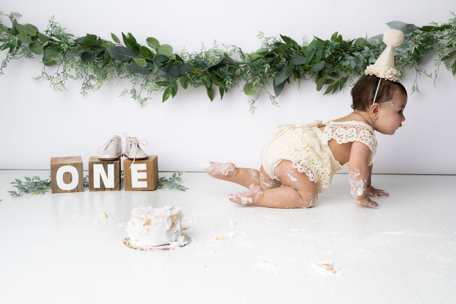 Baby in white lace romper crawls near wooden ONE blocks and smash cake under greenery garland decoration.