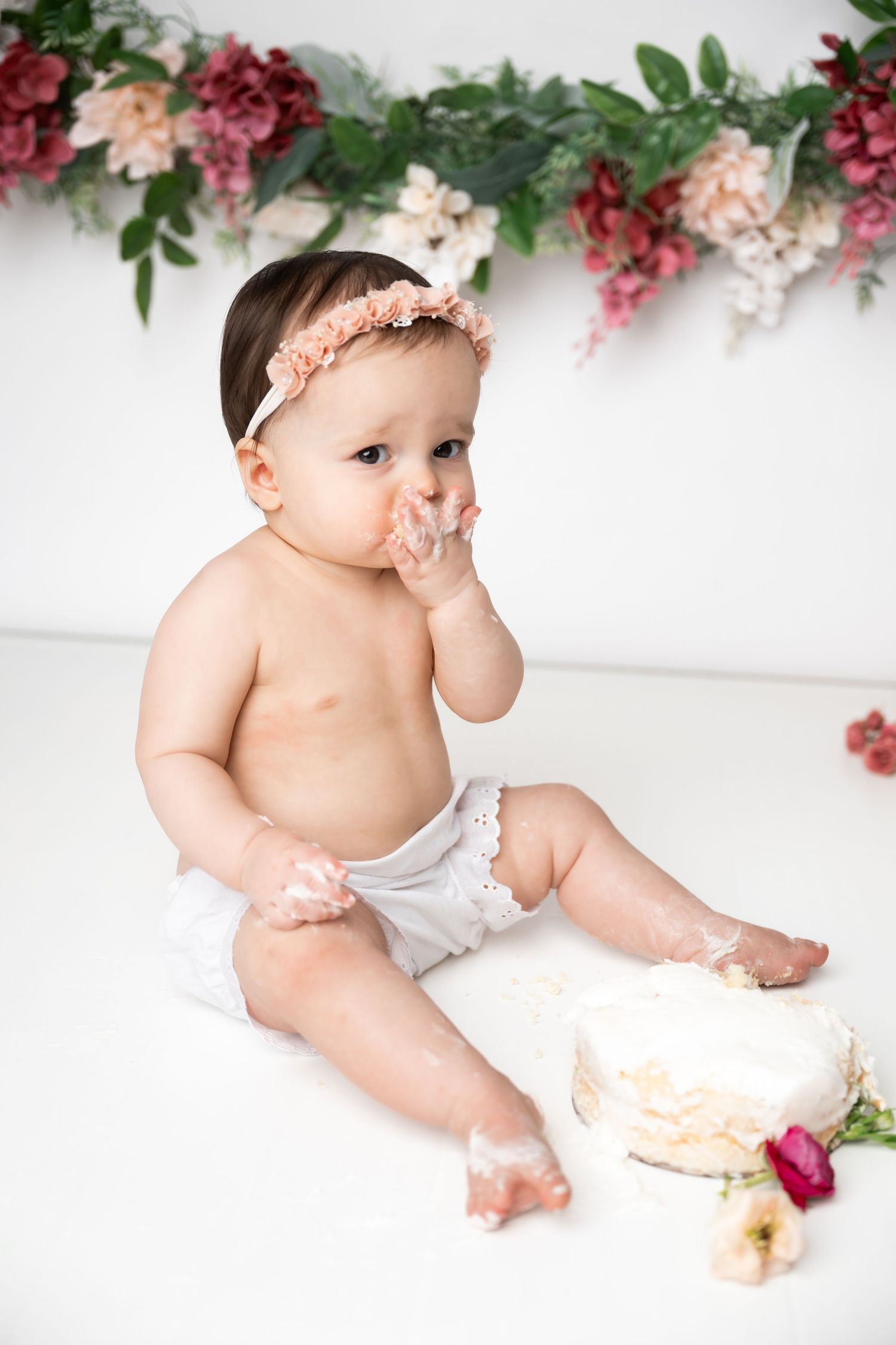 Infant in white diaper wearing peach headband sits on white backdrop with floral garland and cake decorations.