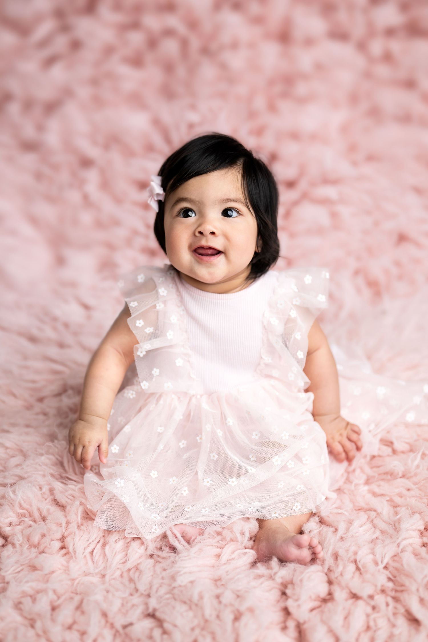 Happy baby in white dress sitting on pink fur backdrop.