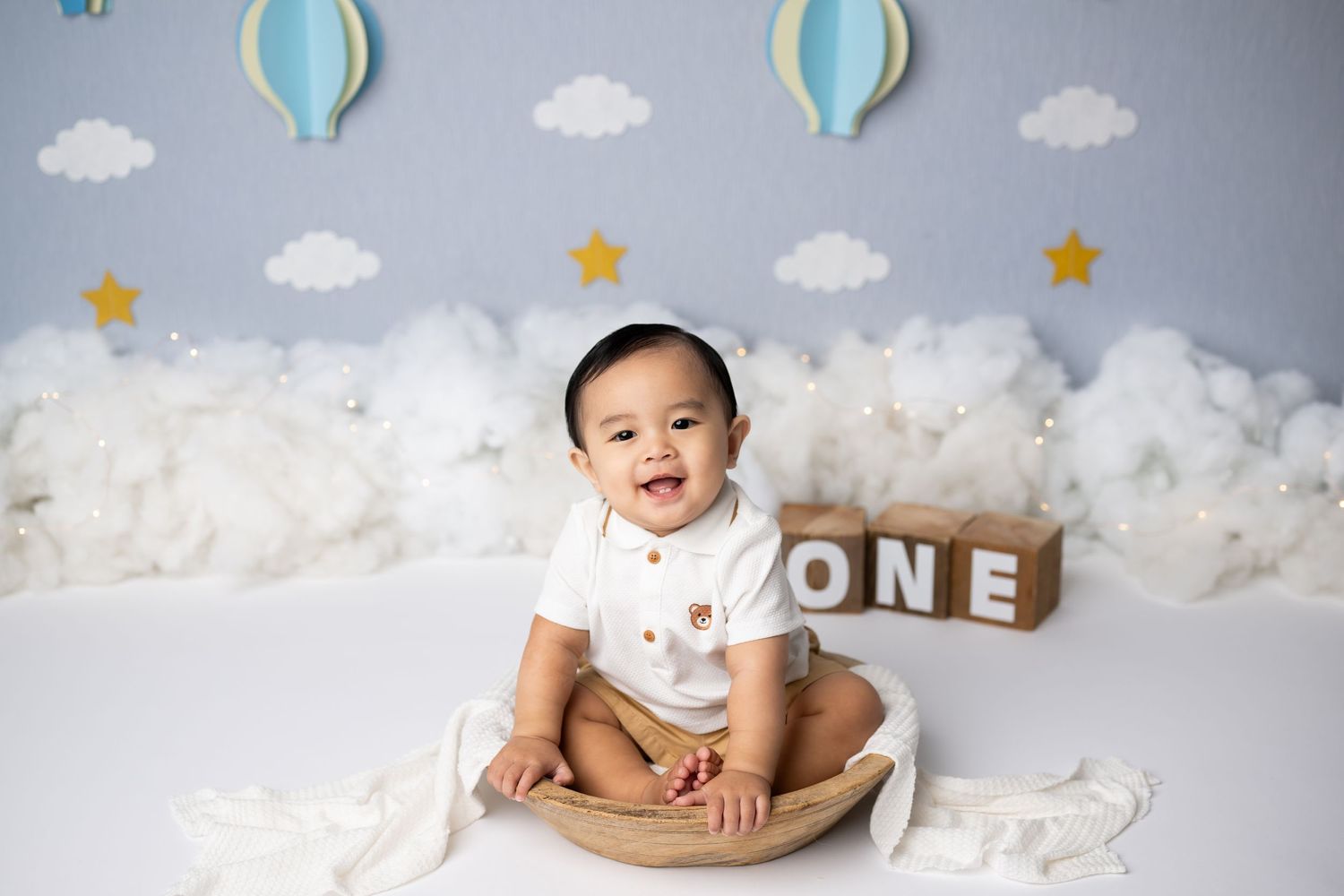 First birthday photo setup with hot air balloon decorations, clouds, and wooden blocks spelling 'ONE' on white backdrop.