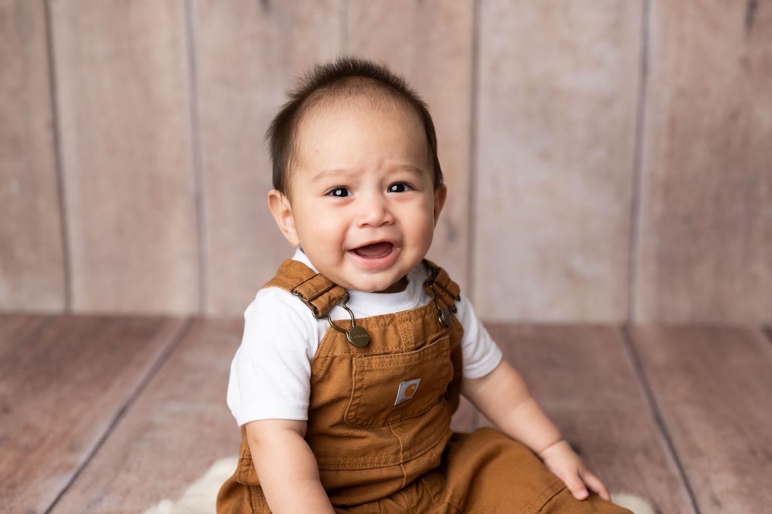 A joyful baby wearing brown overalls and white shirt sits on wooden floor while smiling brightly.