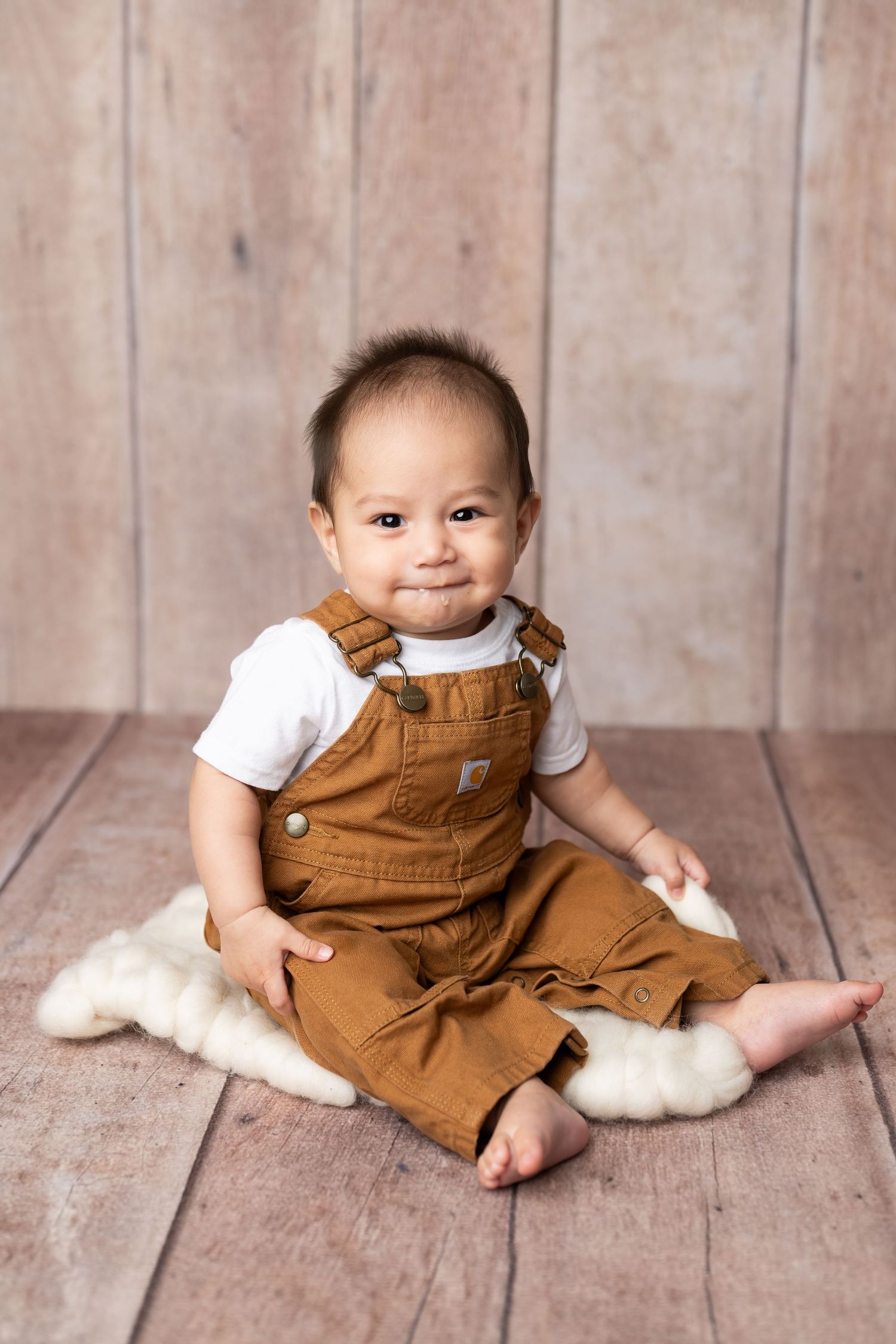 Small baby wearing brown overalls and white shirt sits on wooden floor with soft white fur underneath.