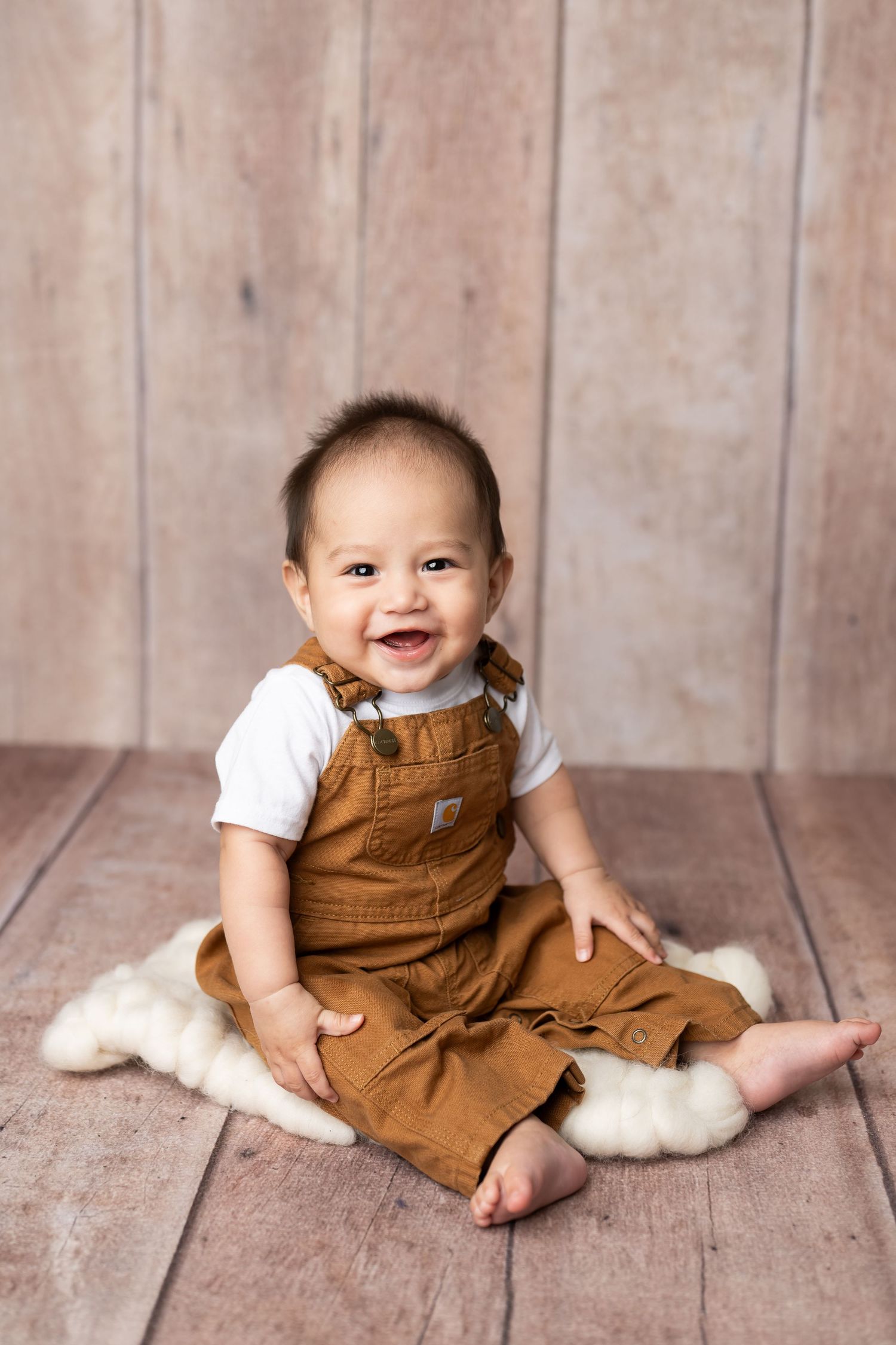 Baby wearing brown overalls sits smiling on white fur rug against wooden backdrop.